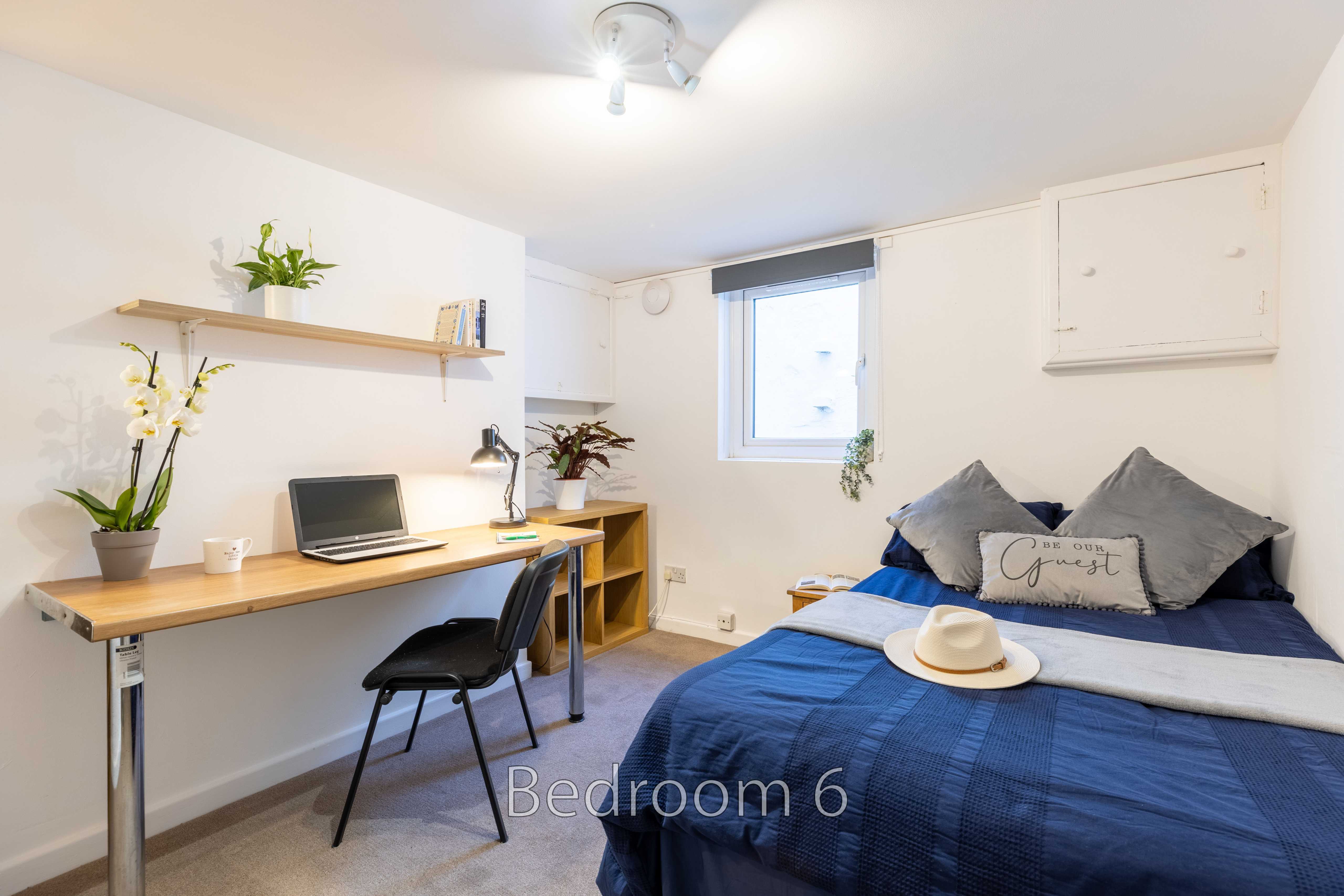 A furnished bedroom with a bed, desk, chair, shelves, window and wall storage.