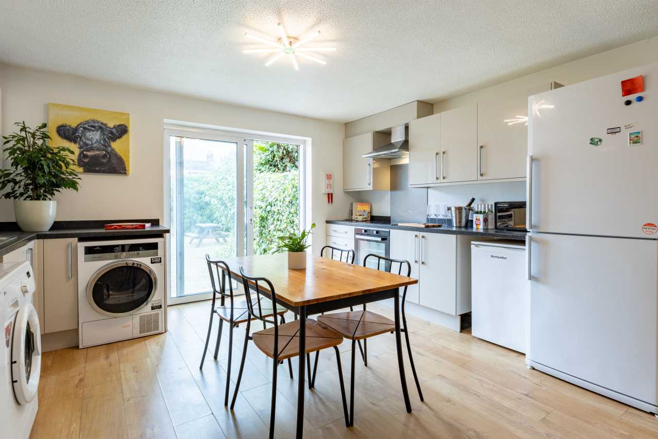 Bright shared kitchen and dining area with white cabinets, appliances, a table and chairs, and glass doors opening to greenery.