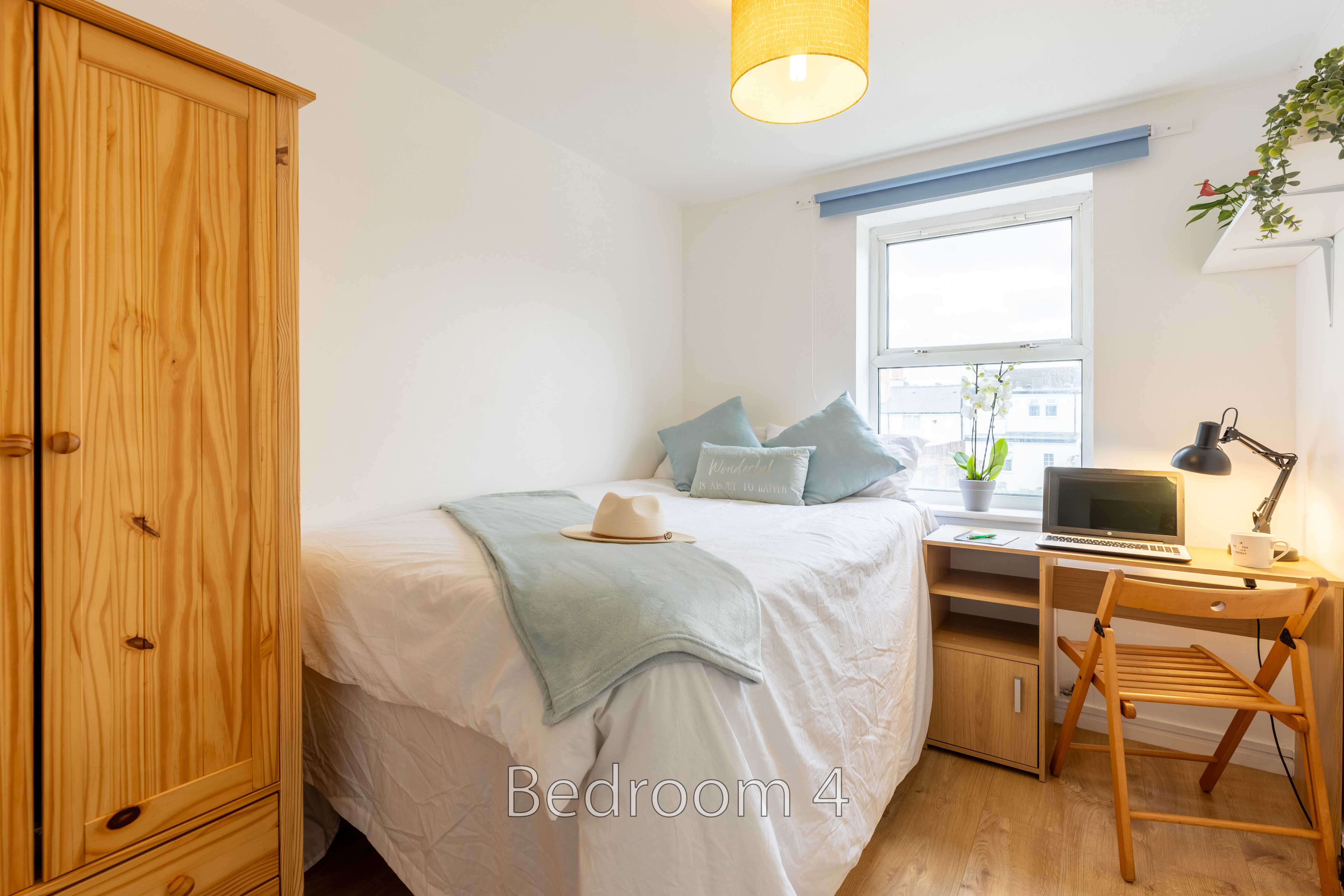 A bright bedroom with a double bed, wooden wardrobe, desk, chair, window and bedside table.