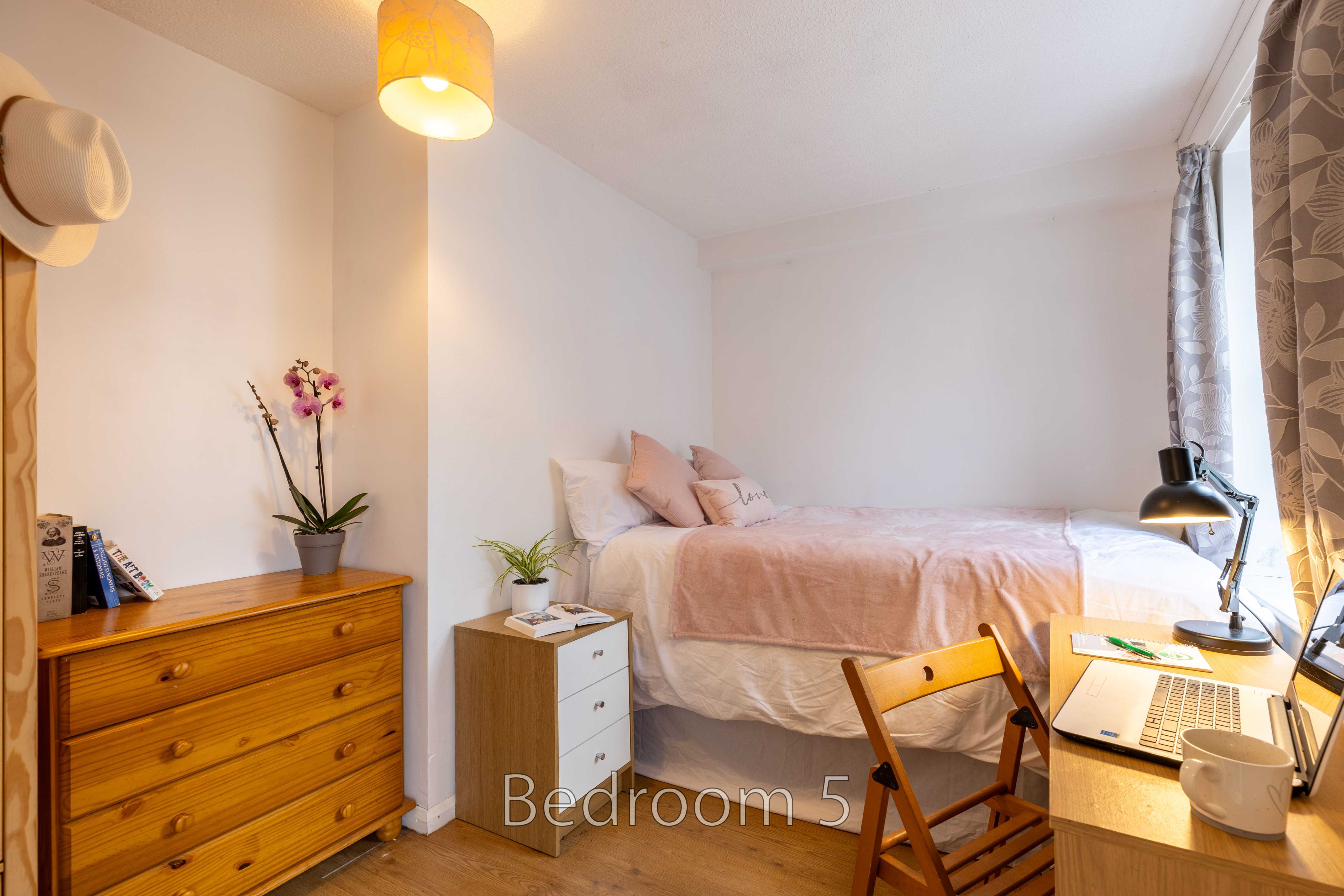A furnished bedroom with a bed, dresser, desk, chair, lamp and window.