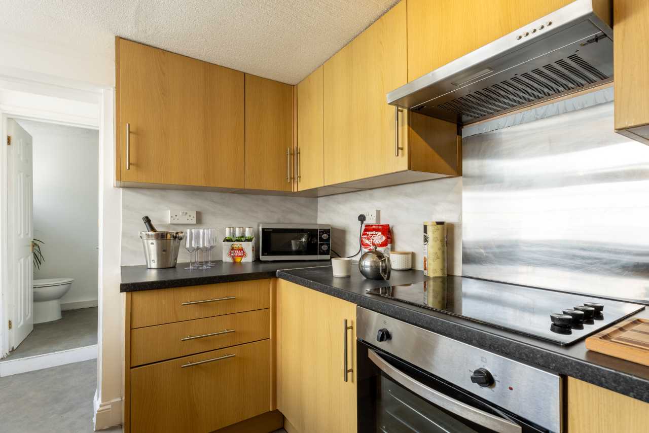 A compact kitchen with wooden cabinets, black countertops, a microwave, hob and oven. An open doorway leads to a bathroom.