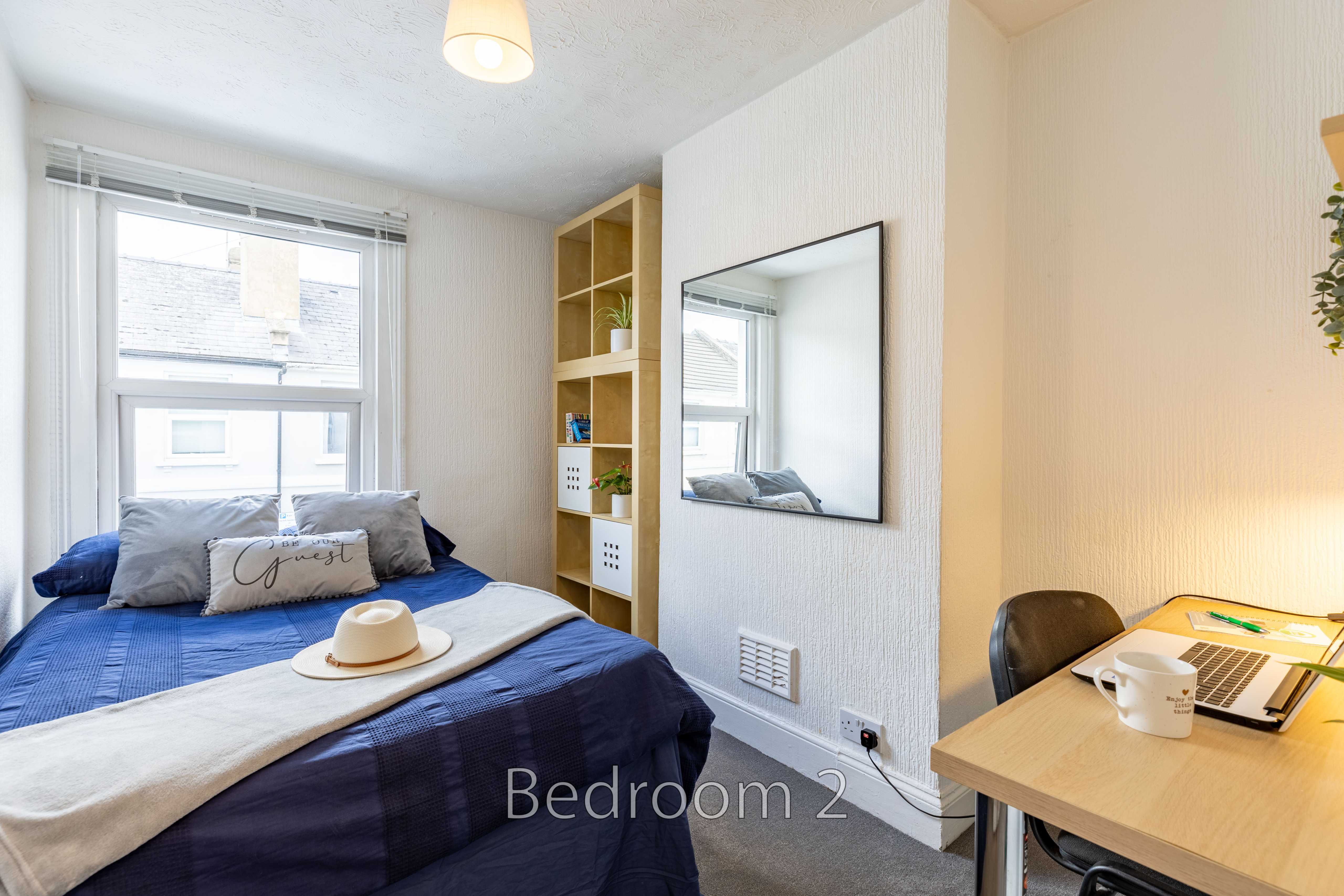 A furnished bedroom with a double bed, window, shelving, mirror and desk.