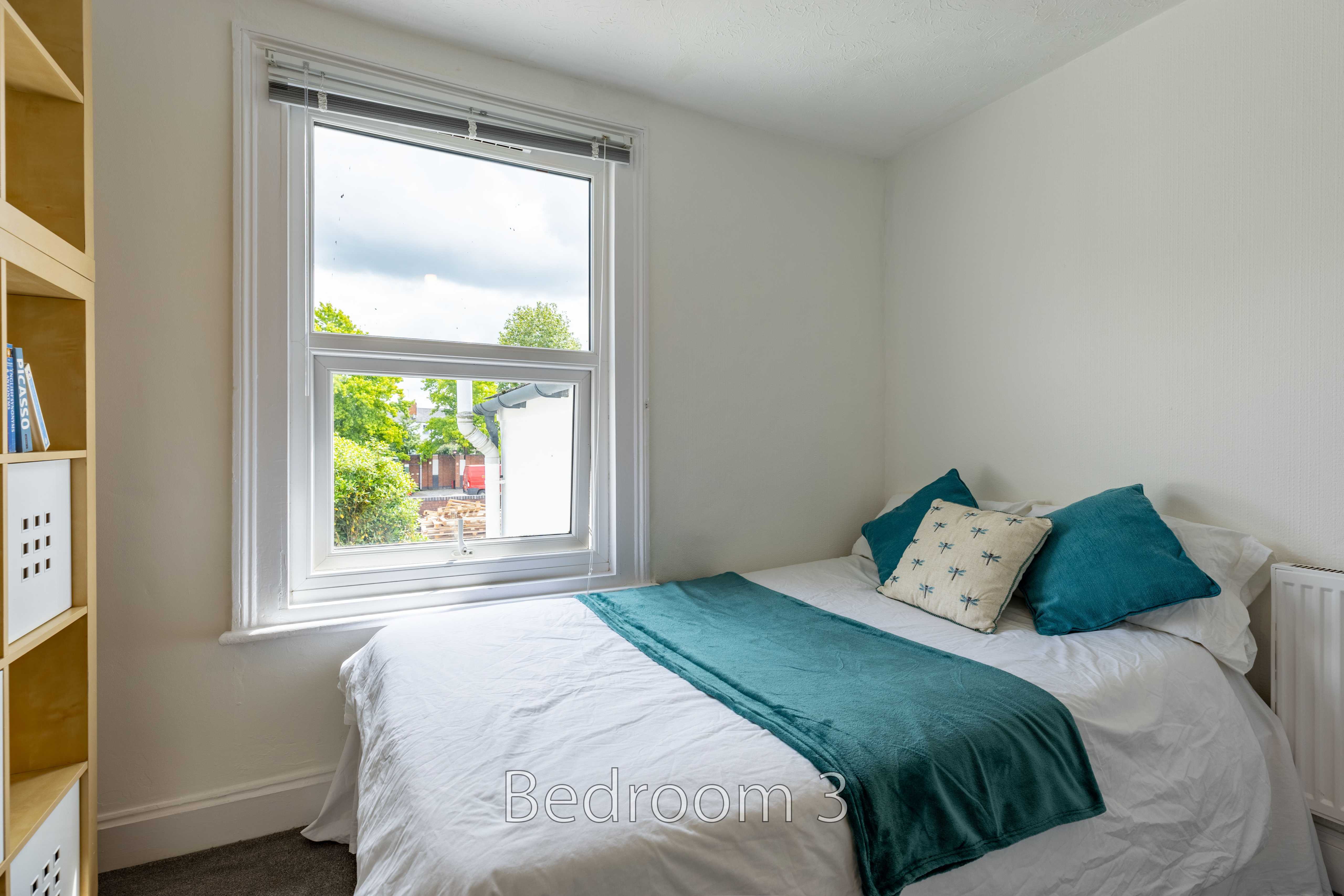 A bedroom with a double bed, teal bedding, a window, shelving and a radiator.