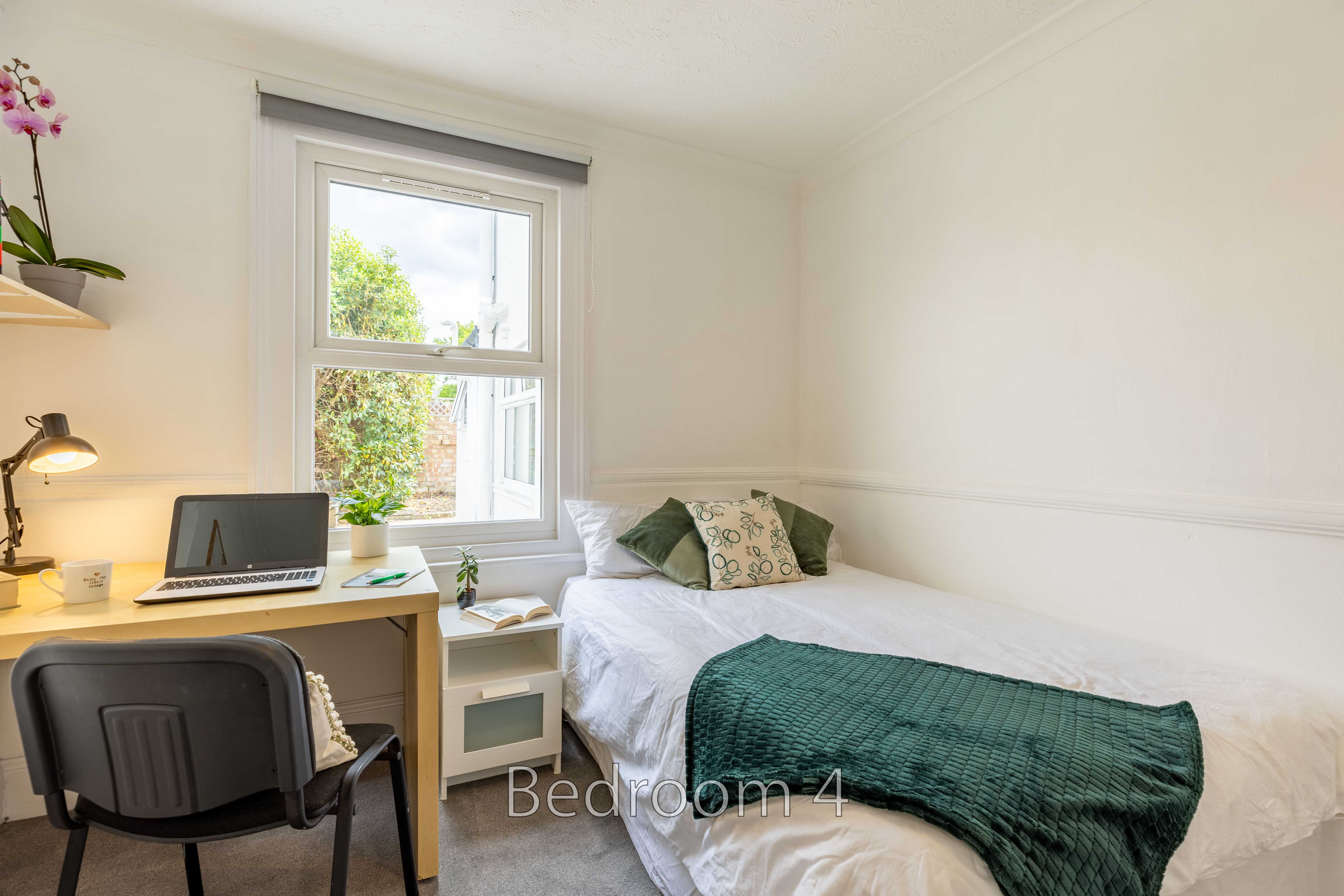 A bright single bedroom with a bed, desk, chair, window and green blanket.