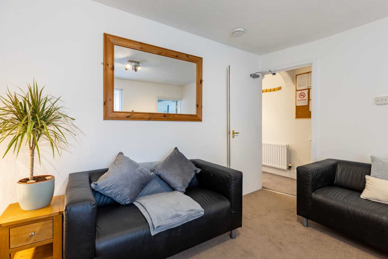 Shared living room with black sofas, cushions, a wooden-framed mirror, a side table and a potted plant.