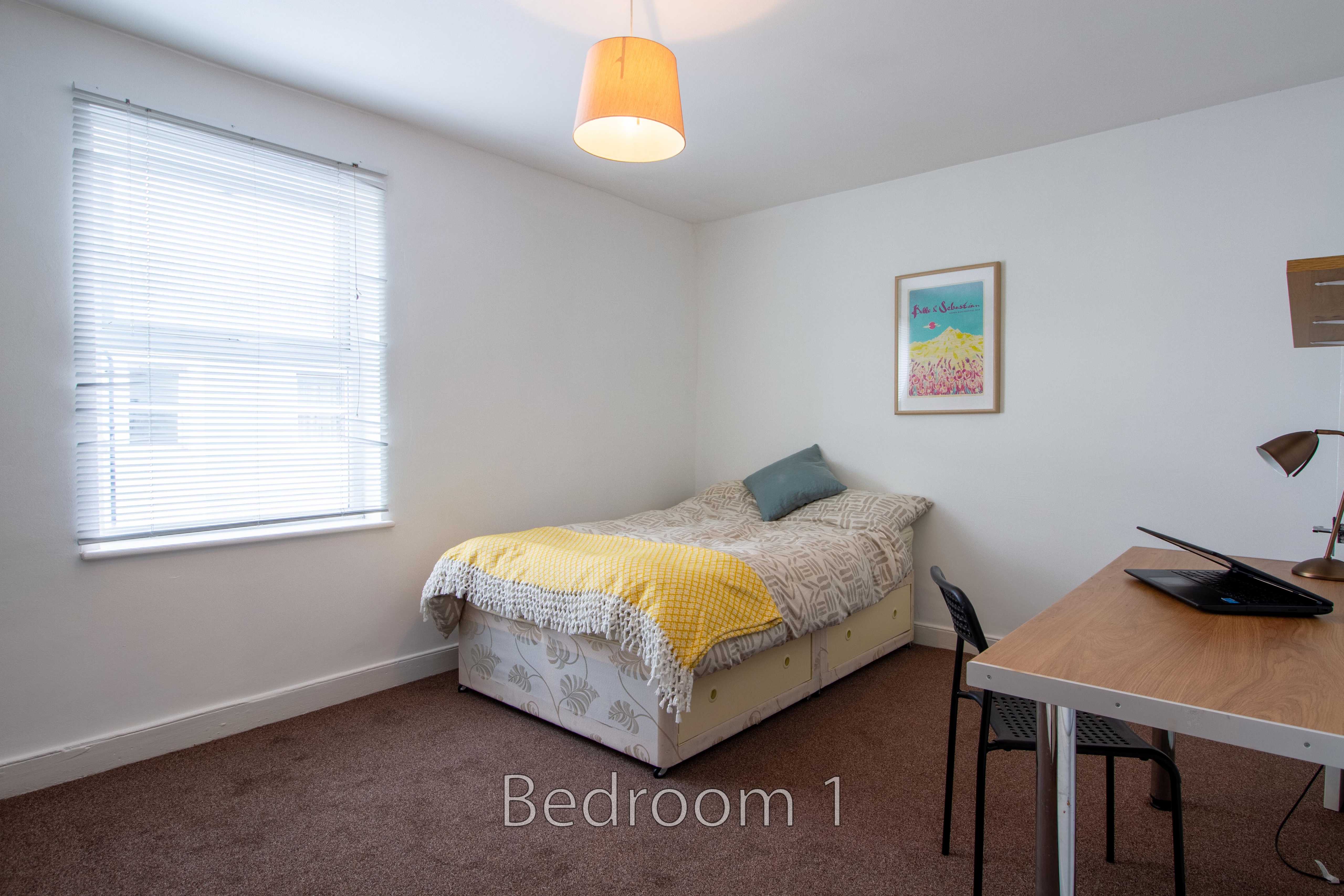 A bedroom with a single bed, desk, chair, window with blinds, and carpeted floor.