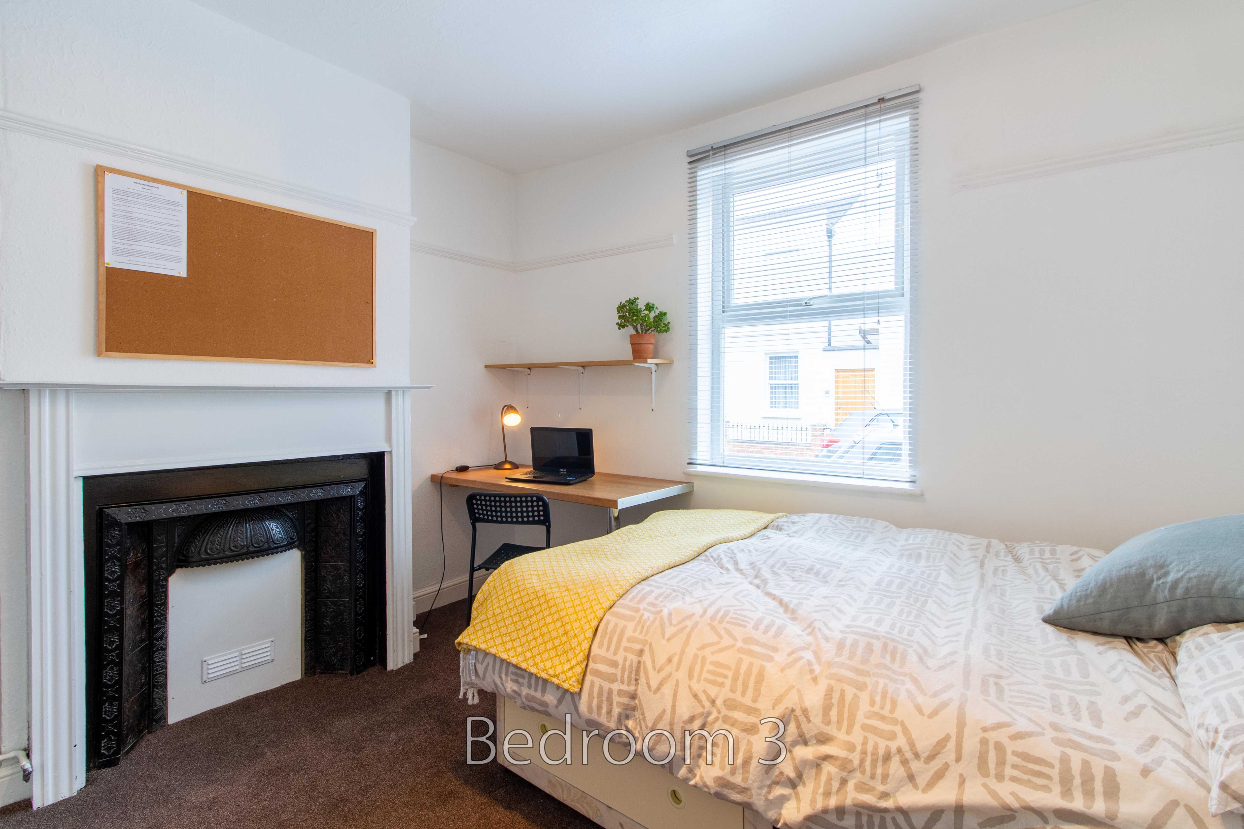 A bedroom with a bed, fireplace, desk, window and wall shelves.