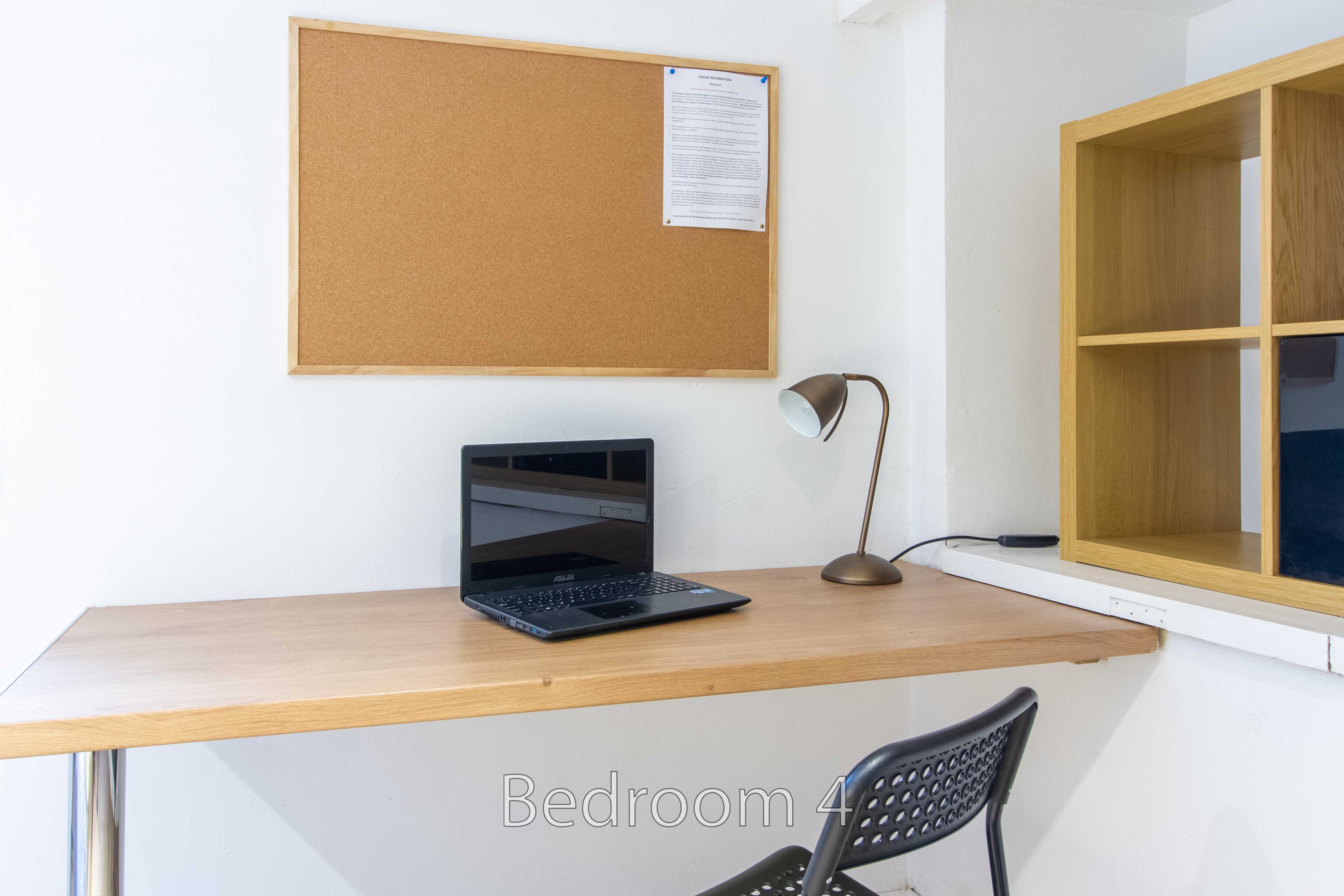 A bedroom workspace with a wooden desk, laptop, lamp, chair, shelving and noticeboard.