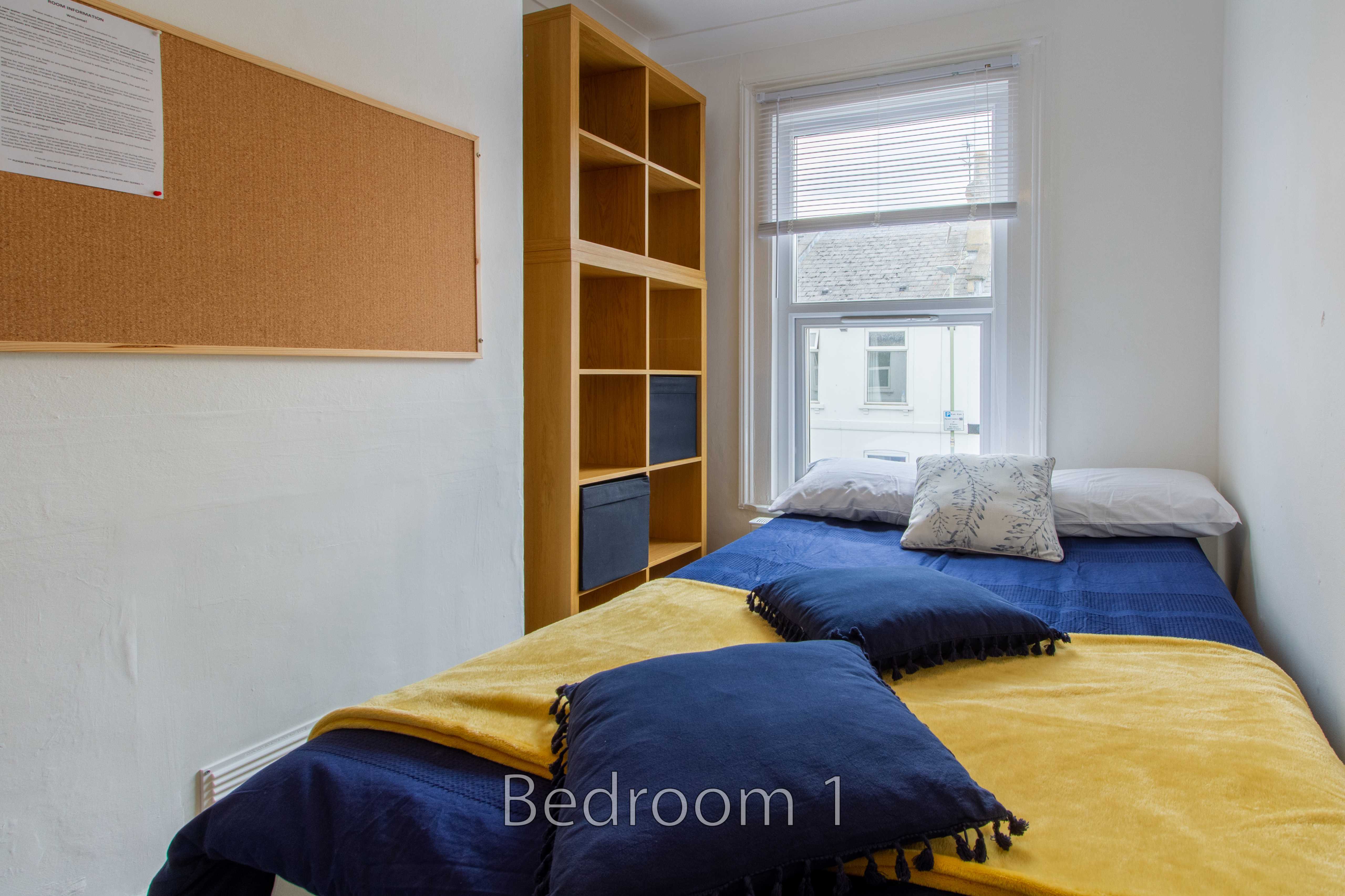 A bedroom with two single beds, colorful blankets, a wooden shelving unit, a corkboard, and a window.