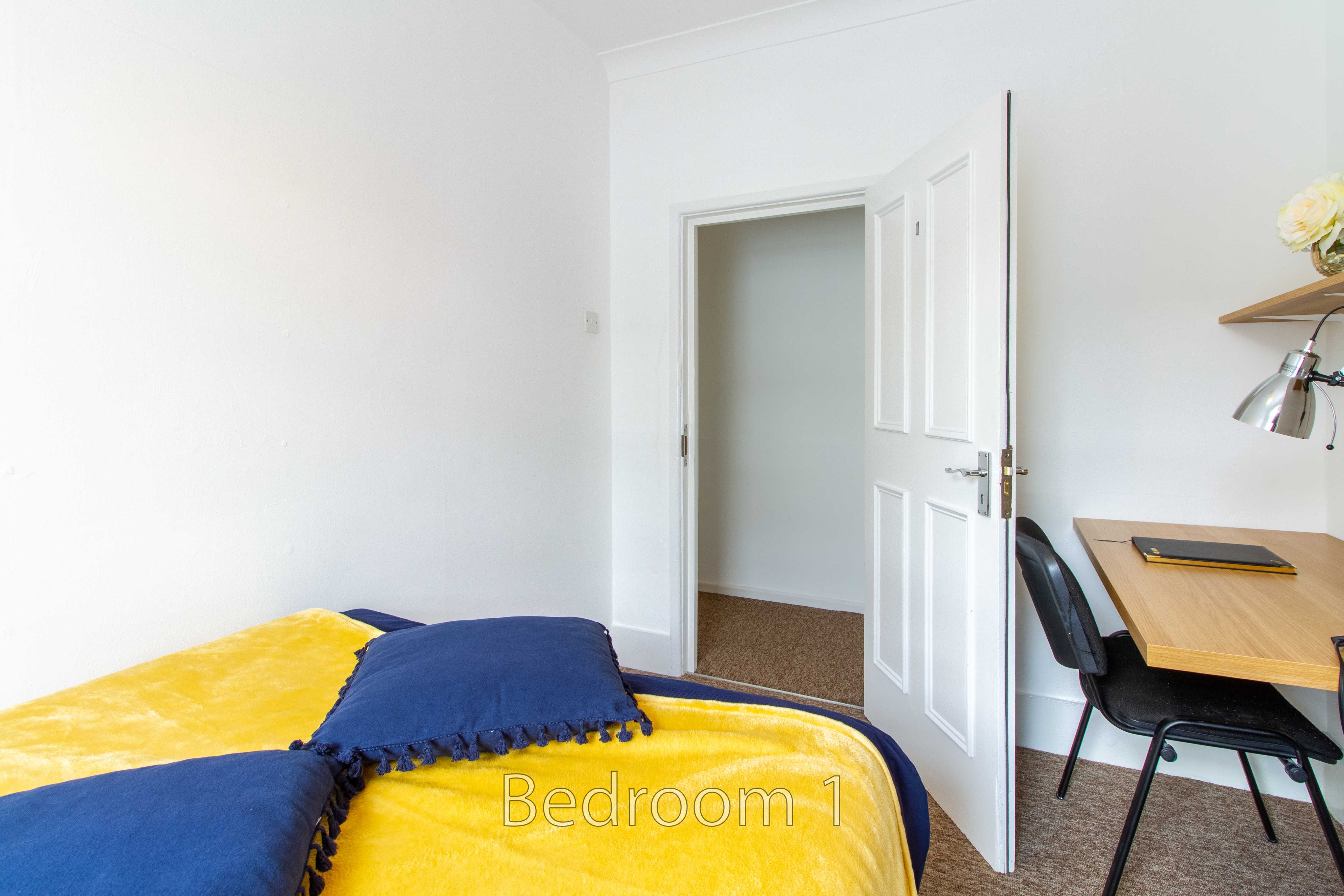A bedroom with a bed covered by a yellow and blue blanket, a desk and chair, and an open door.