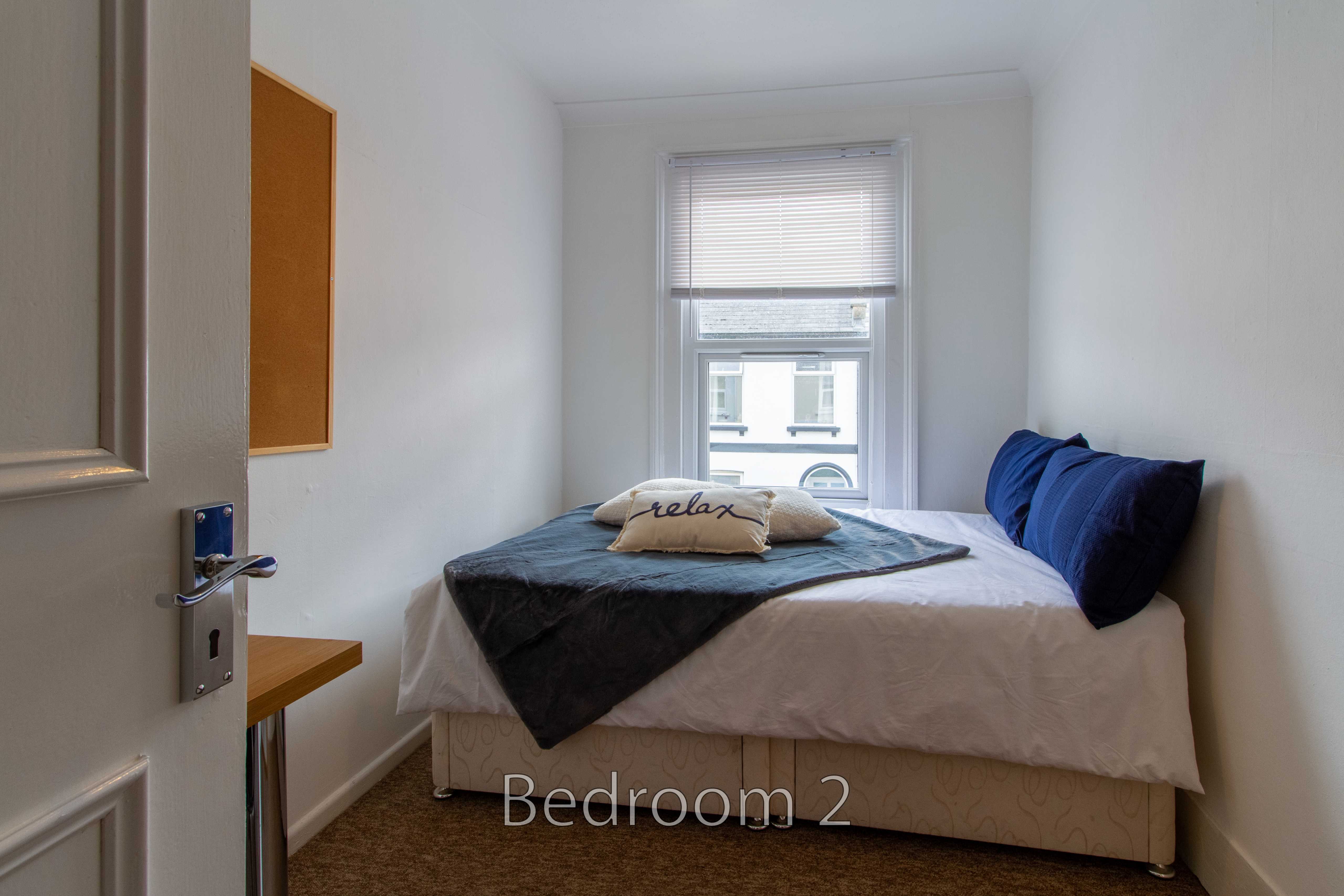 A small bedroom with a double bed, blue cushions, a window with blinds, and a small desk.