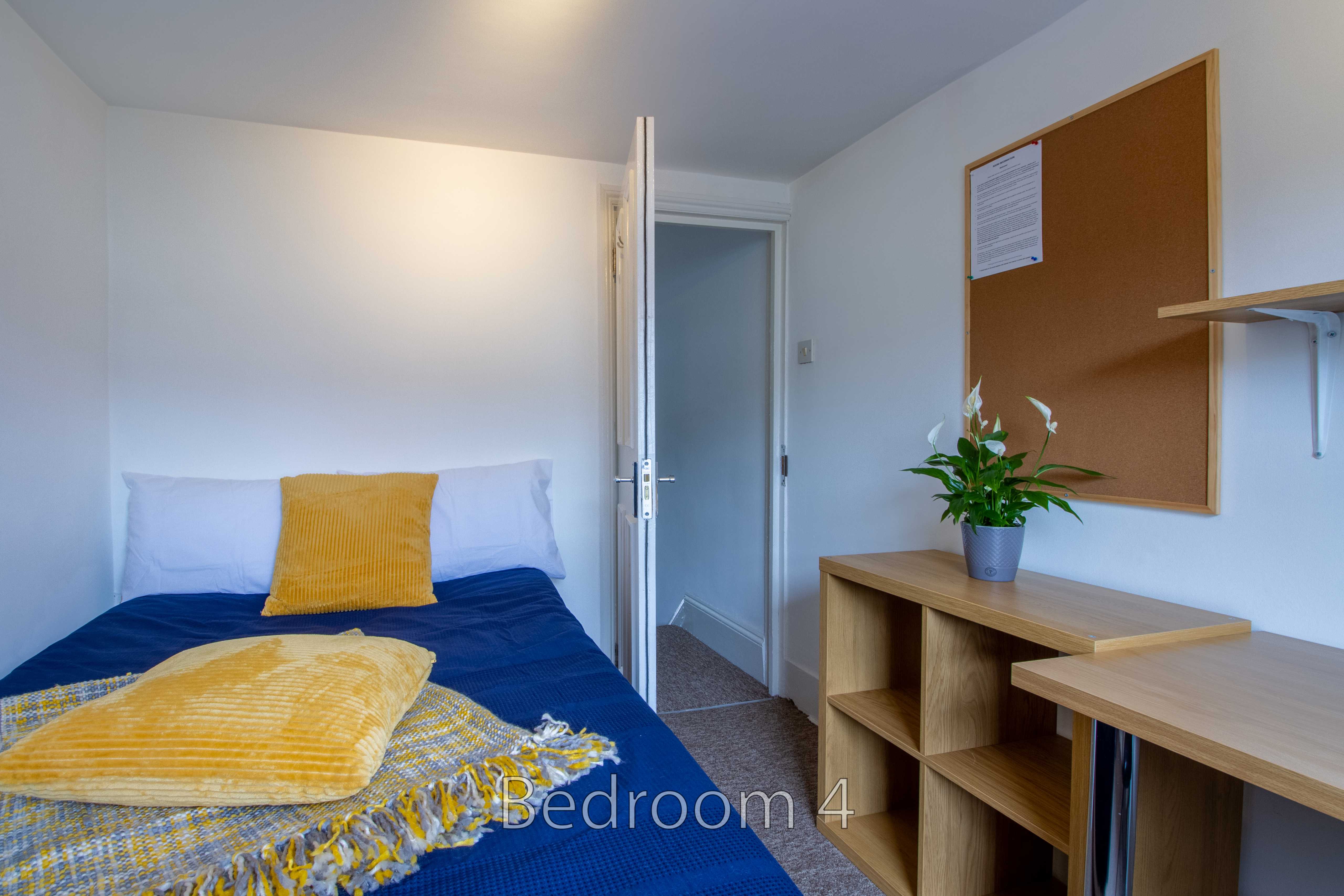 A furnished bedroom with a single bed, desk, shelving unit, bulletin board and potted plant.