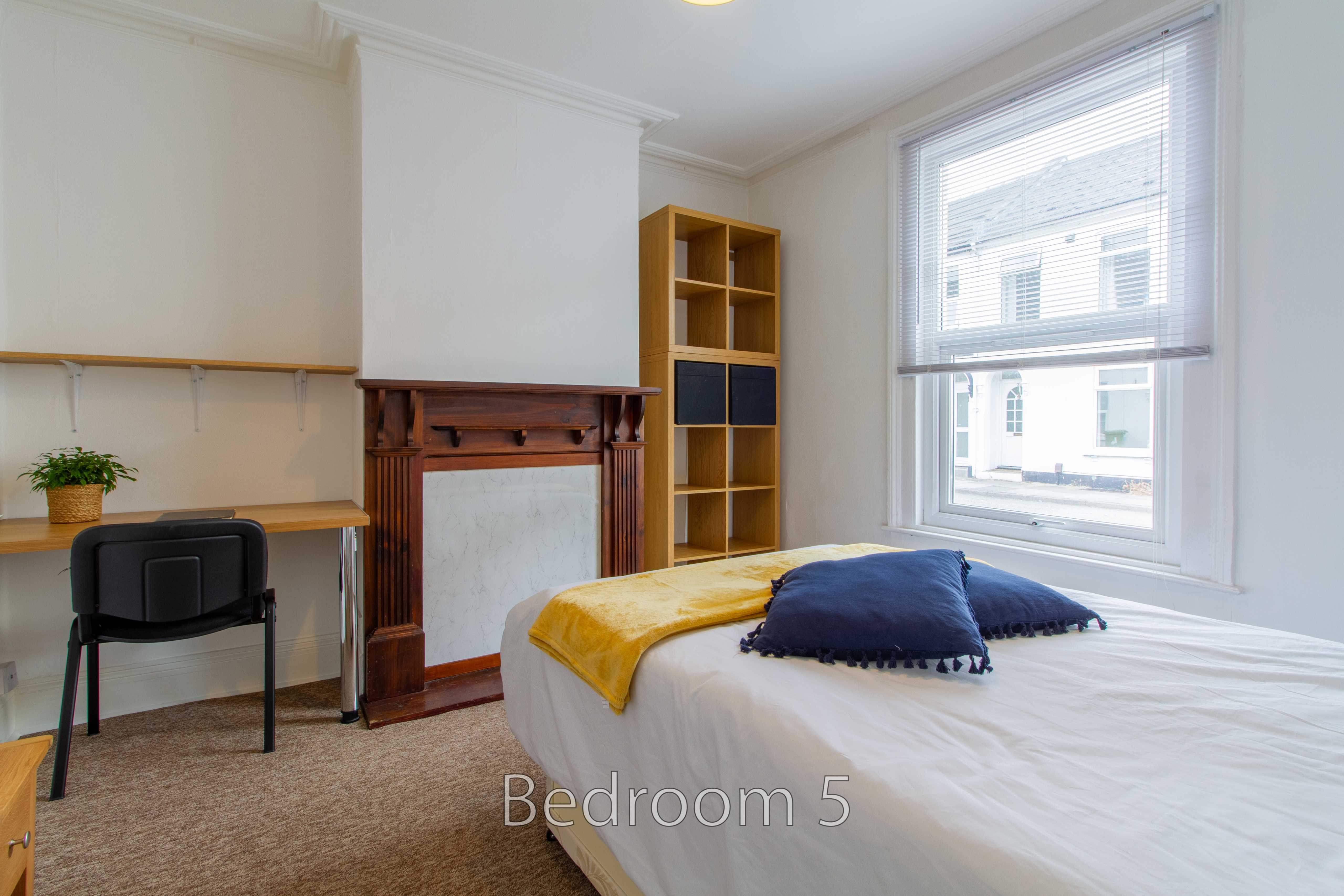 Bedroom with a bed, fireplace, desk, chair, shelving unit and large window.