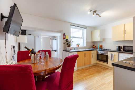 A bright open-plan kitchen and dining area with wooden cabinets, appliances, a dining table and red upholstered chairs.