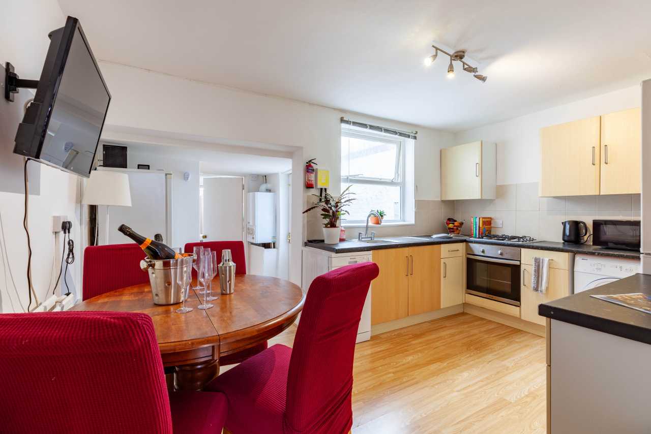 A bright open-plan kitchen and dining area with wooden cabinets, appliances, a dining table and red upholstered chairs.