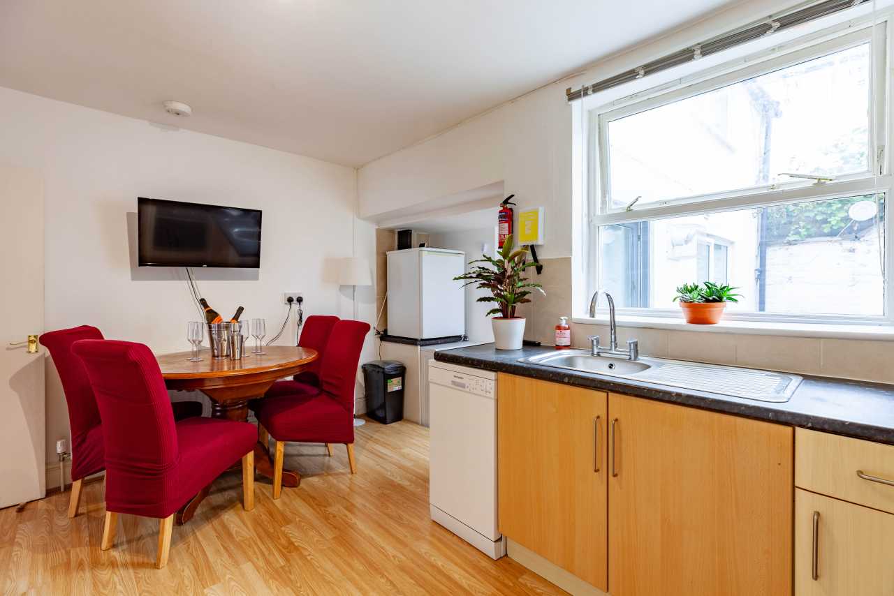A shared kitchen and dining area with wooden cabinets, appliances, a table, red chairs and a large window.