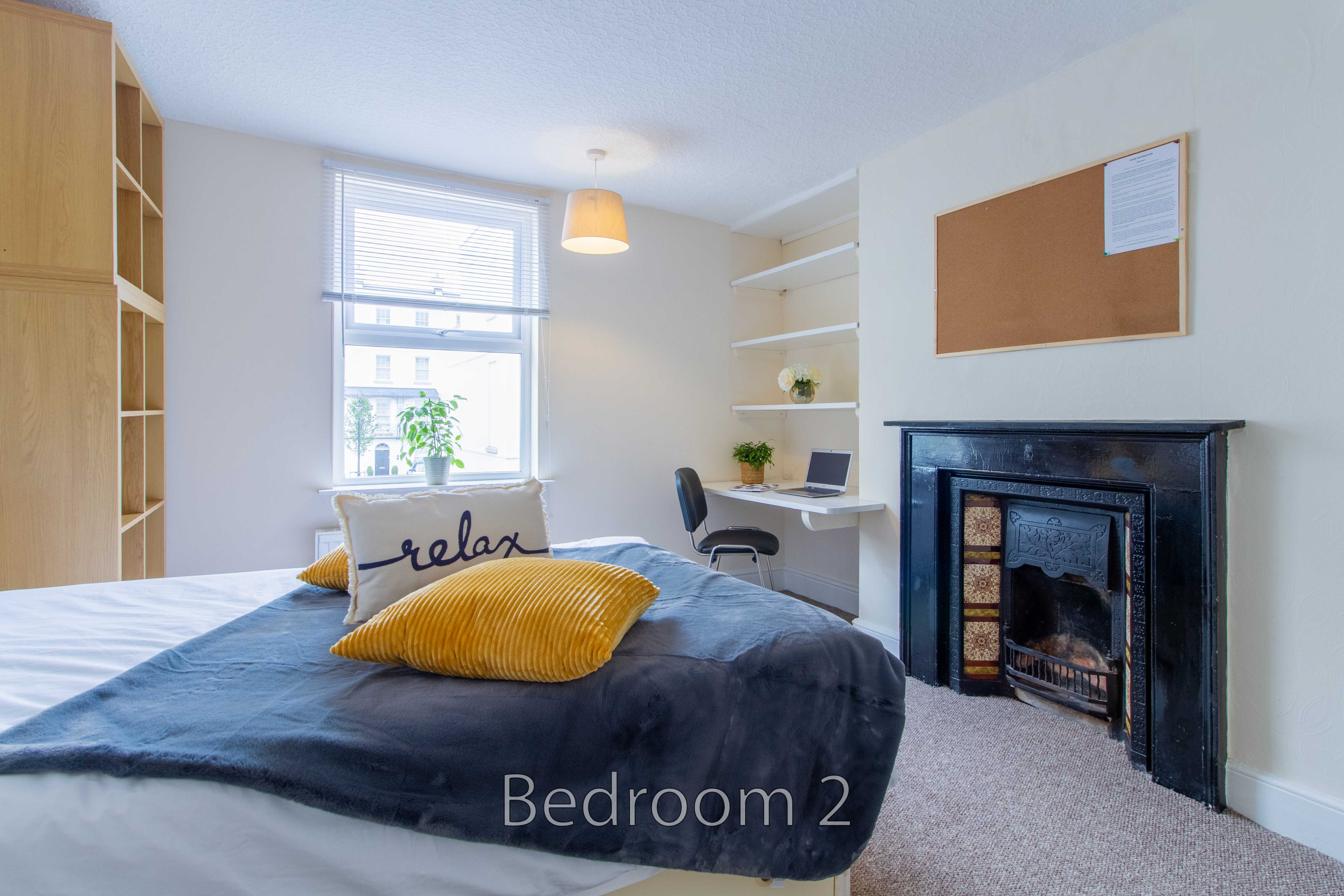 A furnished bedroom with a bed, desk, shelves, window and fireplace.