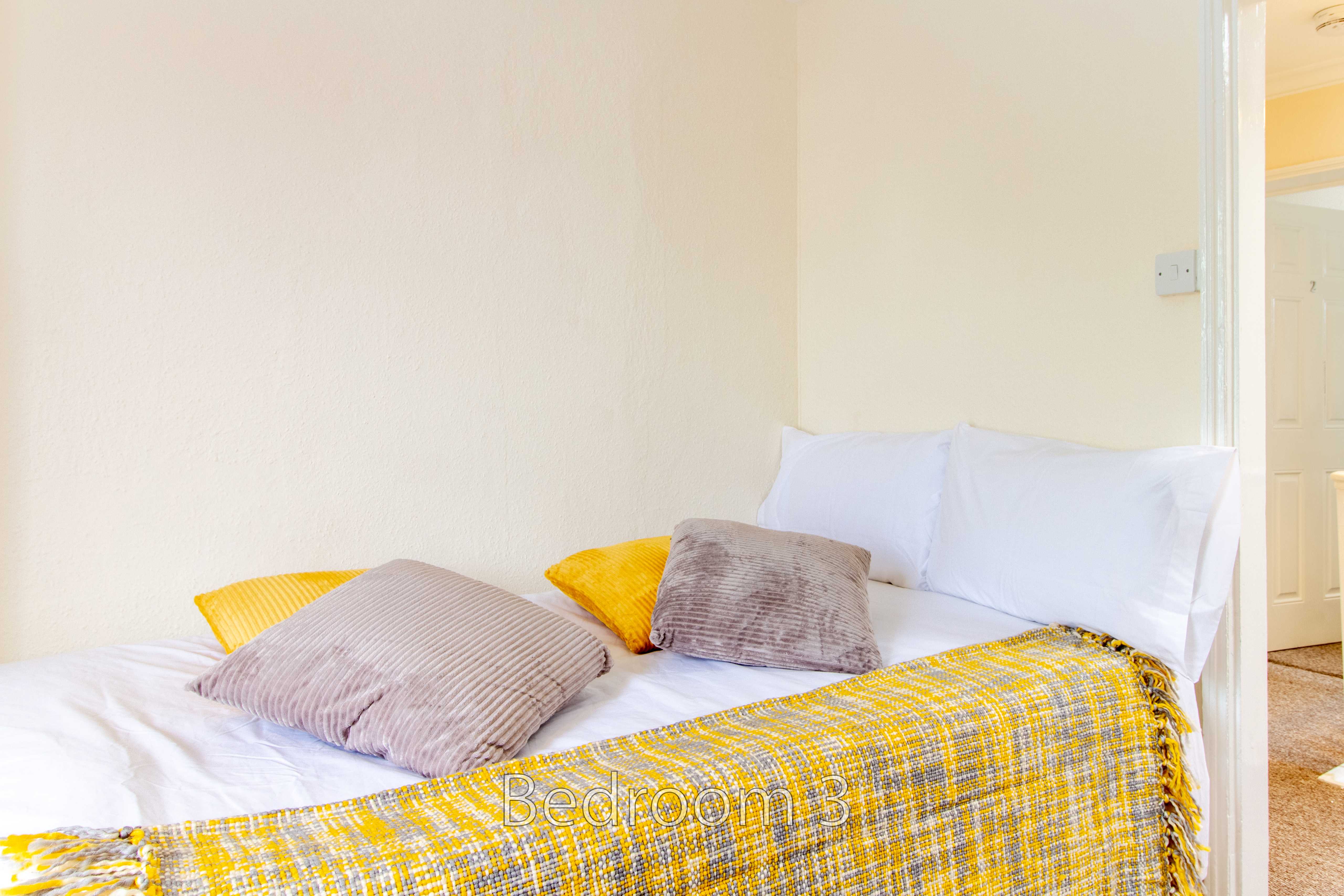 A bedroom with a neatly made bed, white pillows, patterned cushions and a yellow-and-gray throw.
