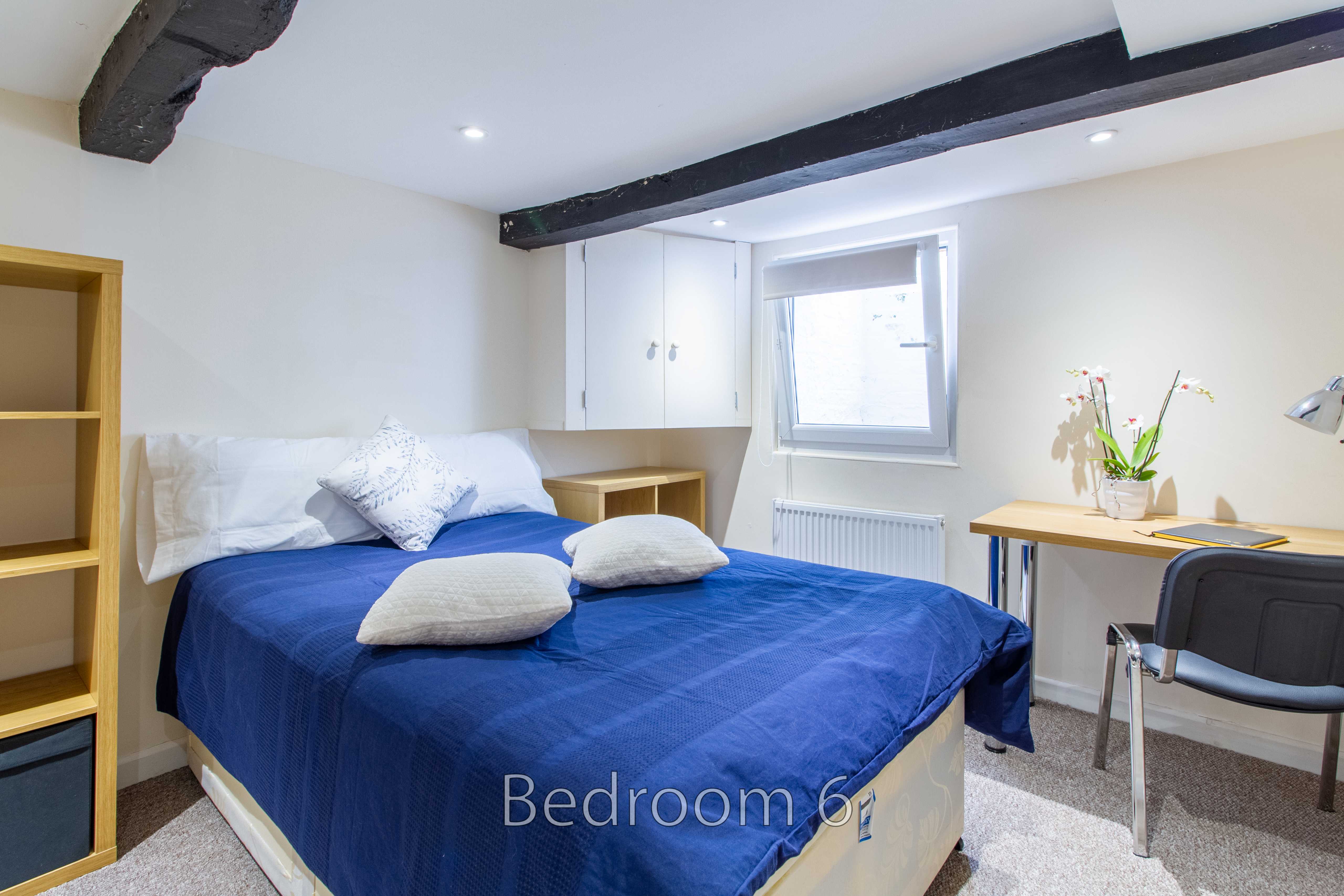 A bedroom with a blue-covered bed, pillows, desk, chair, window and exposed ceiling beams.