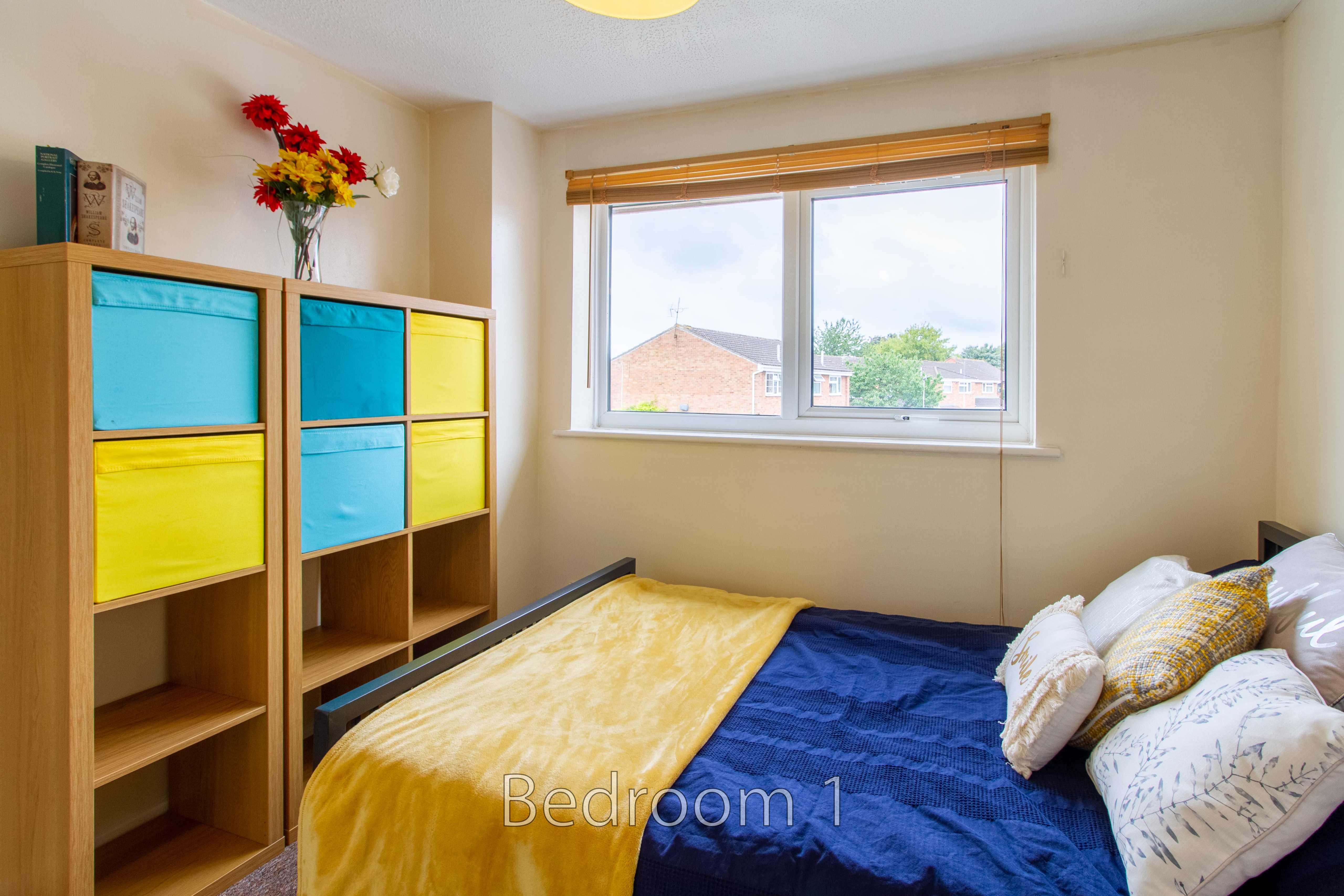 A bedroom with a bed, colorful shelving units, a window, and a vase of flowers.