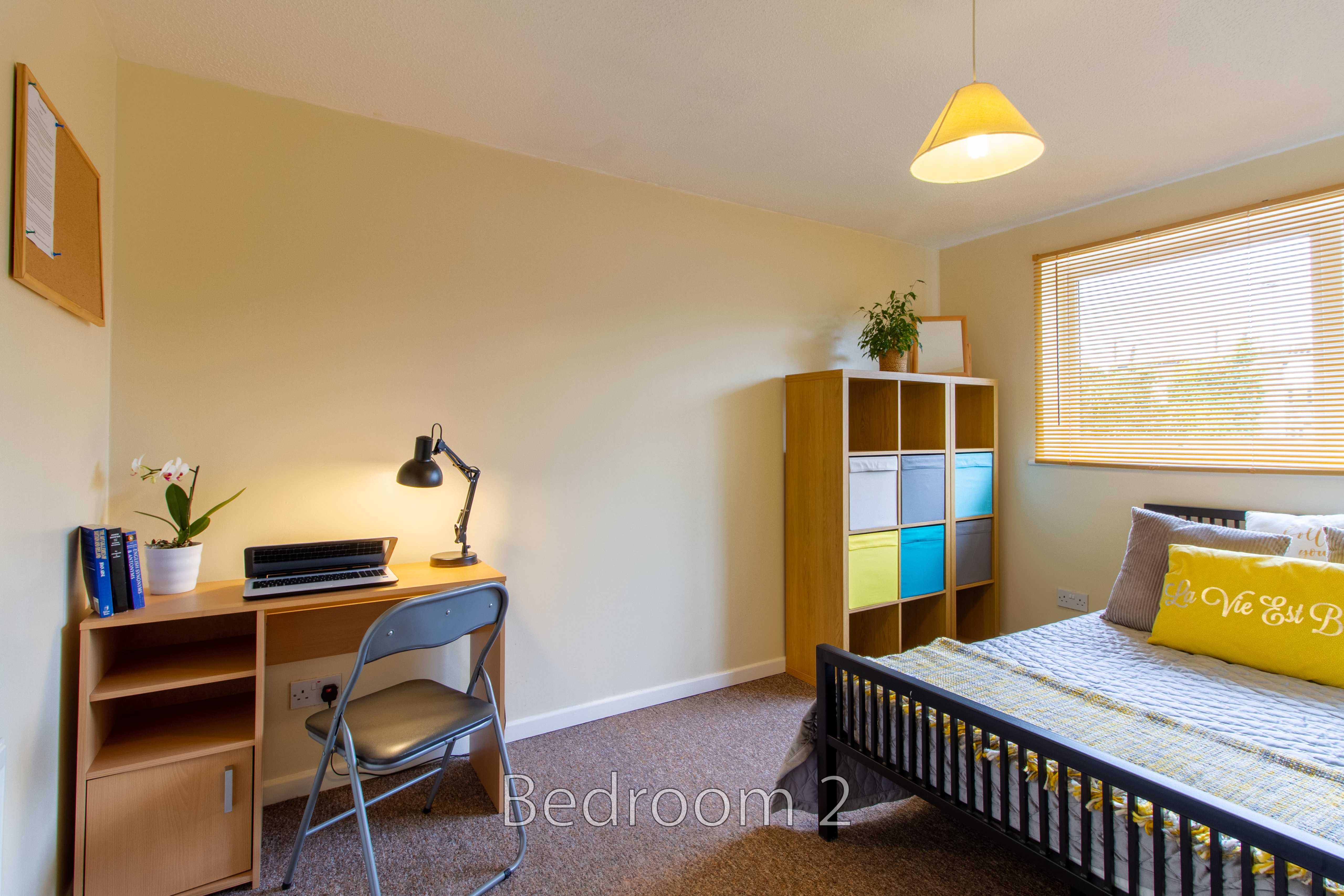 A furnished bedroom with a single bed, desk, chair, shelving unit, lamp and window blinds.