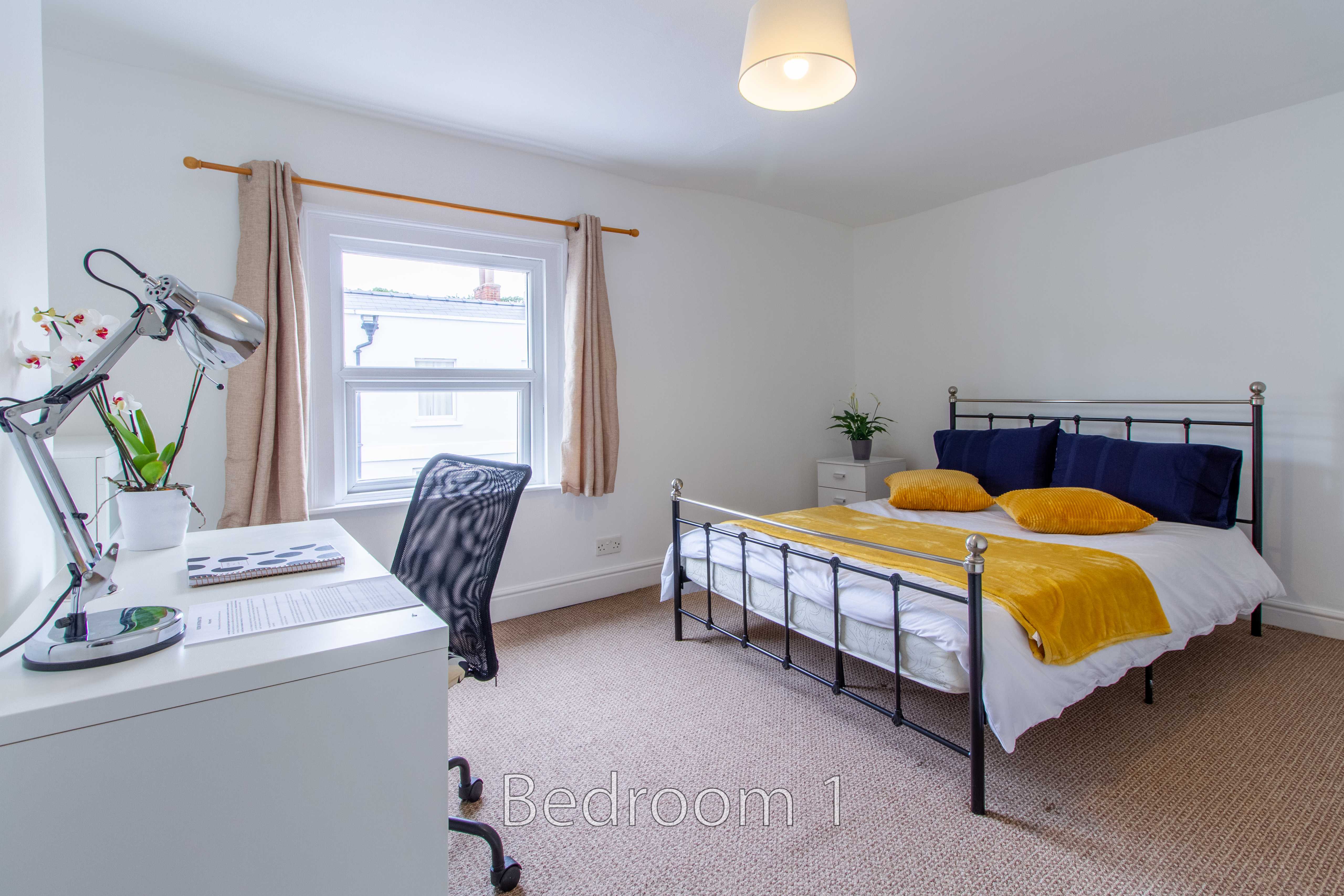 A bedroom with a double bed, yellow bedding, a desk, exercise equipment, and a window.