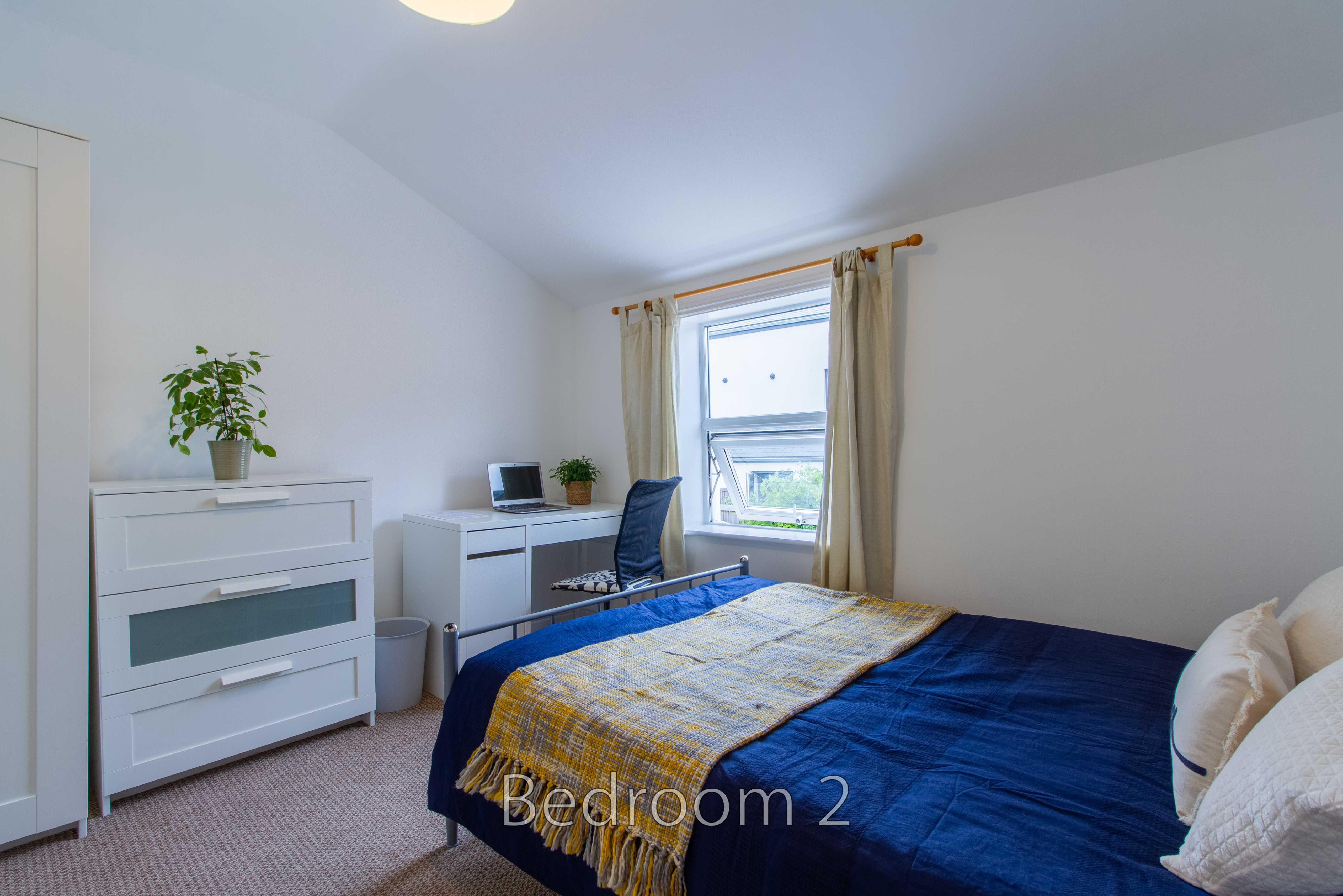 A furnished bedroom with a double bed, chest of drawers, desk, window and curtains.