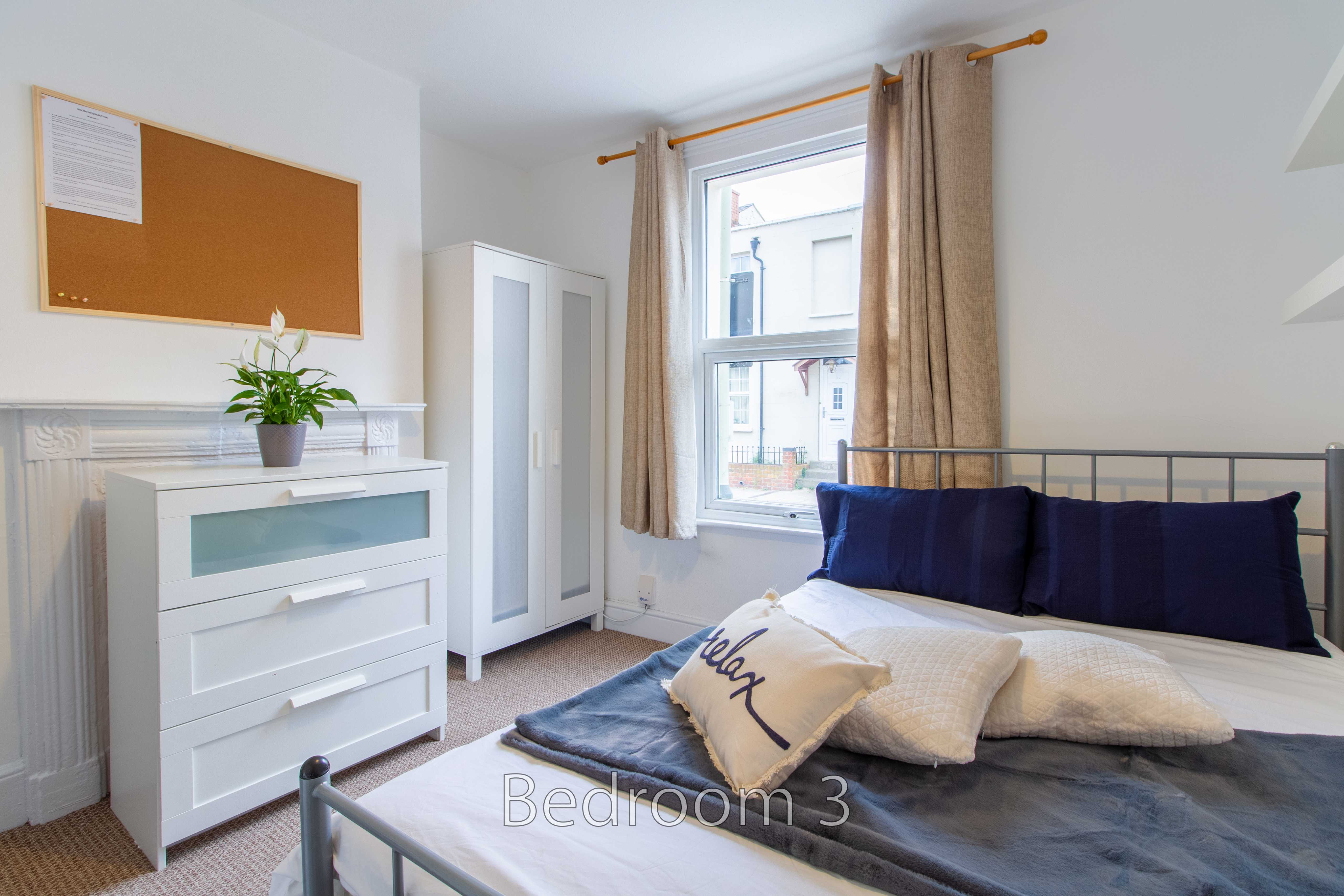 A bright bedroom with a metal-framed bed, bedside storage, a chest of drawers, a wardrobe, curtains and a window.