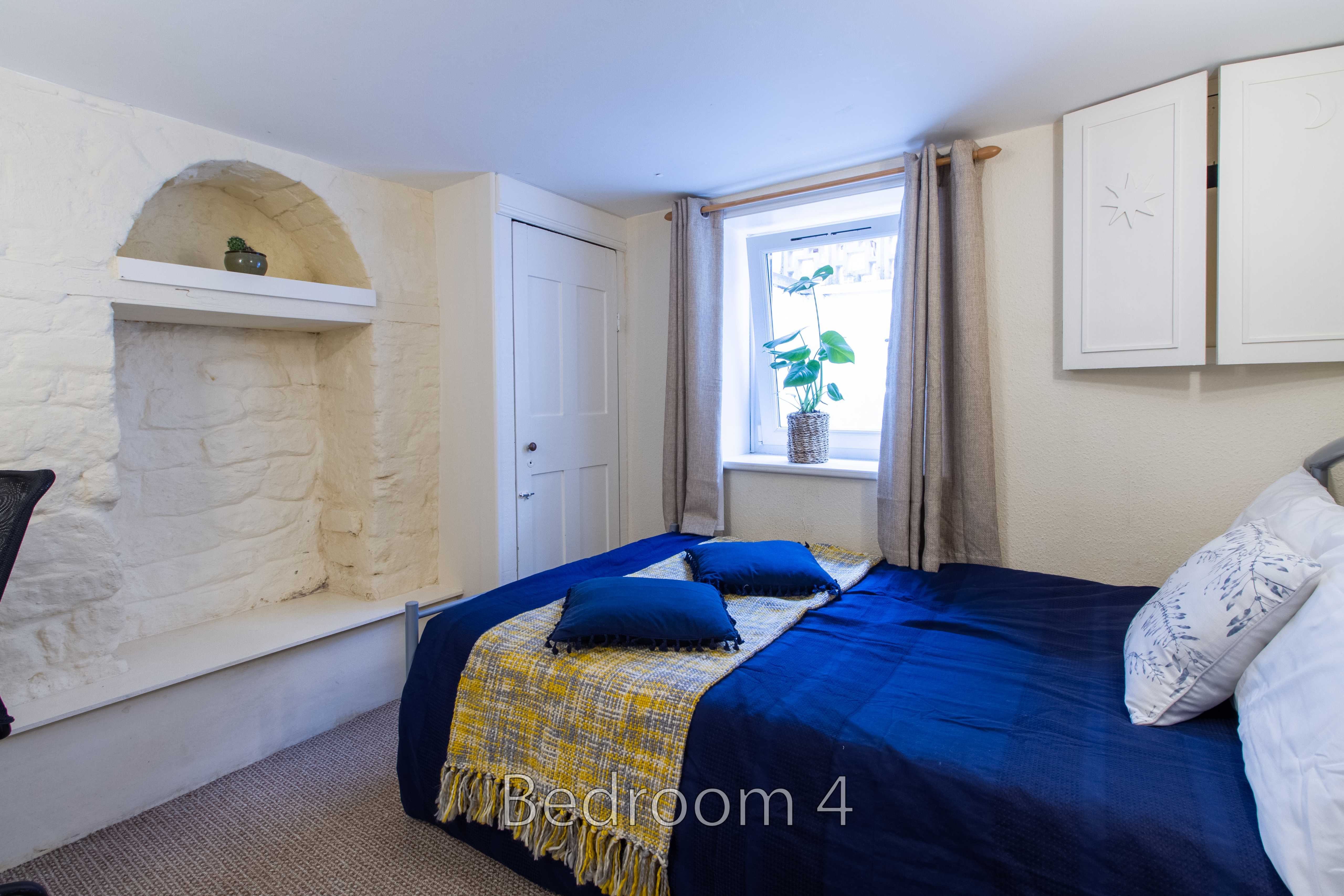 A furnished bedroom with a double bed, blue bedding, a window, built-in cabinets and a white stone fireplace recess.