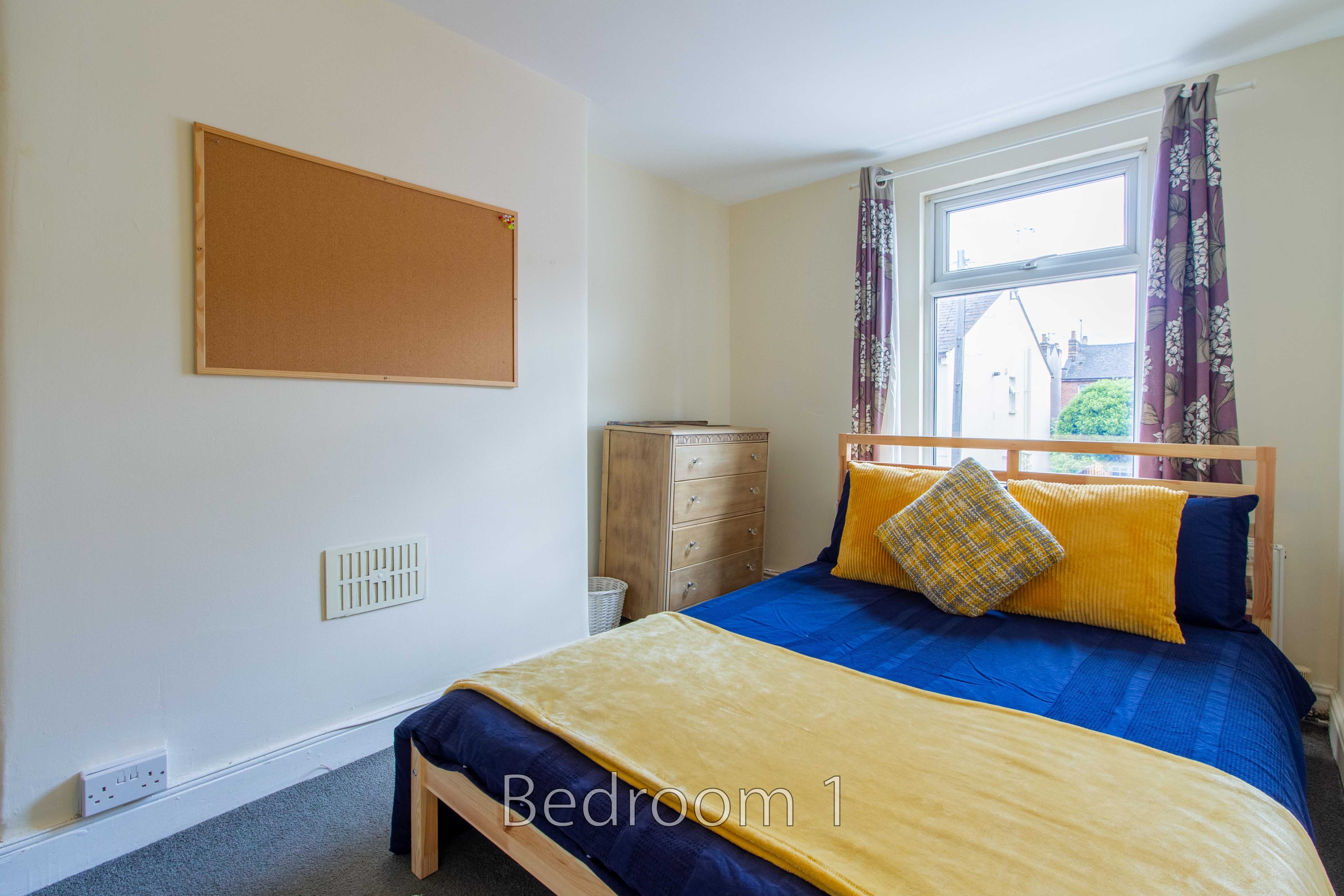 A furnished bedroom with a double bed, chest of drawers, window, curtains and wall-mounted noticeboard.