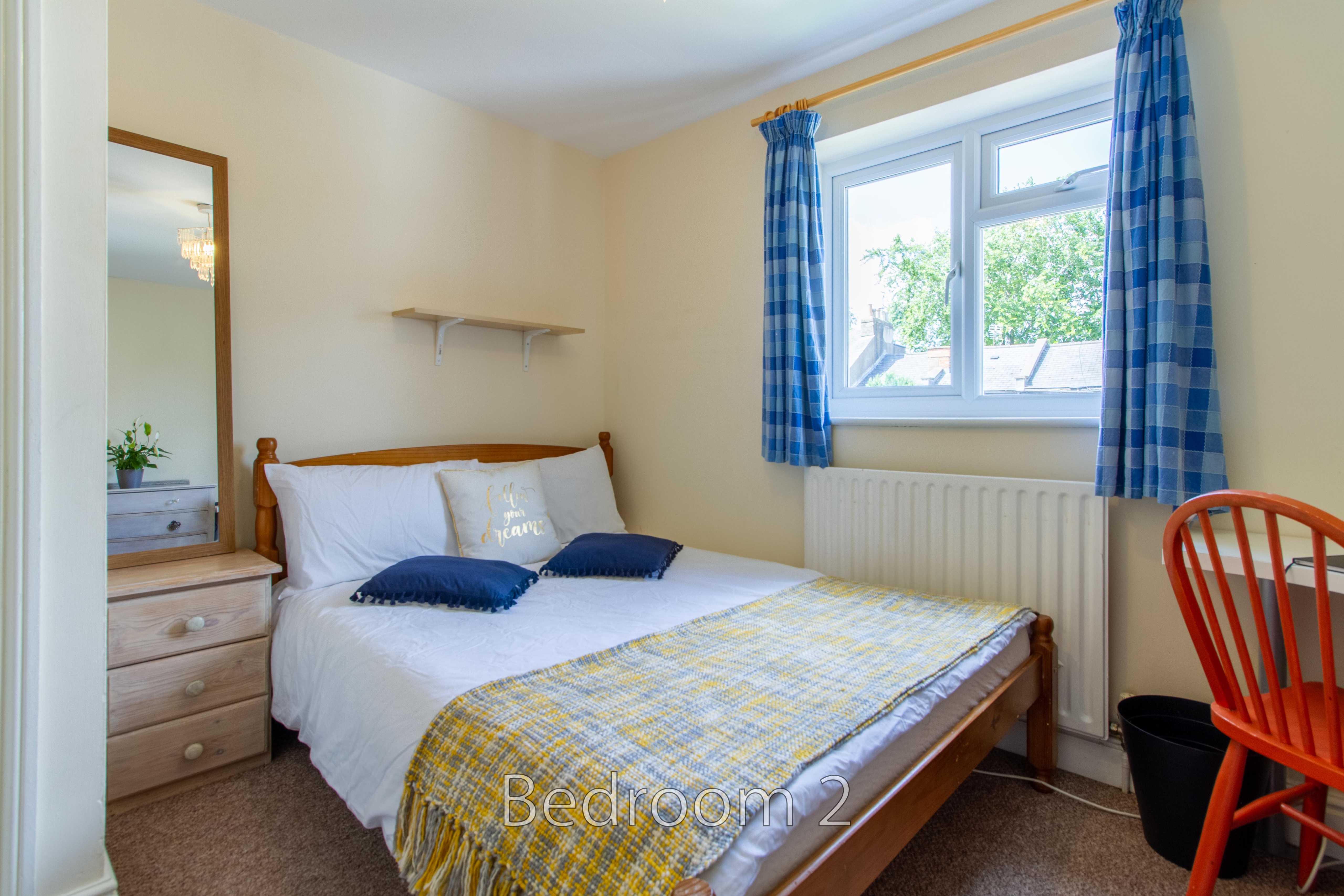 A furnished bedroom with a double bed, dresser, window, radiator, curtains and wooden chair.