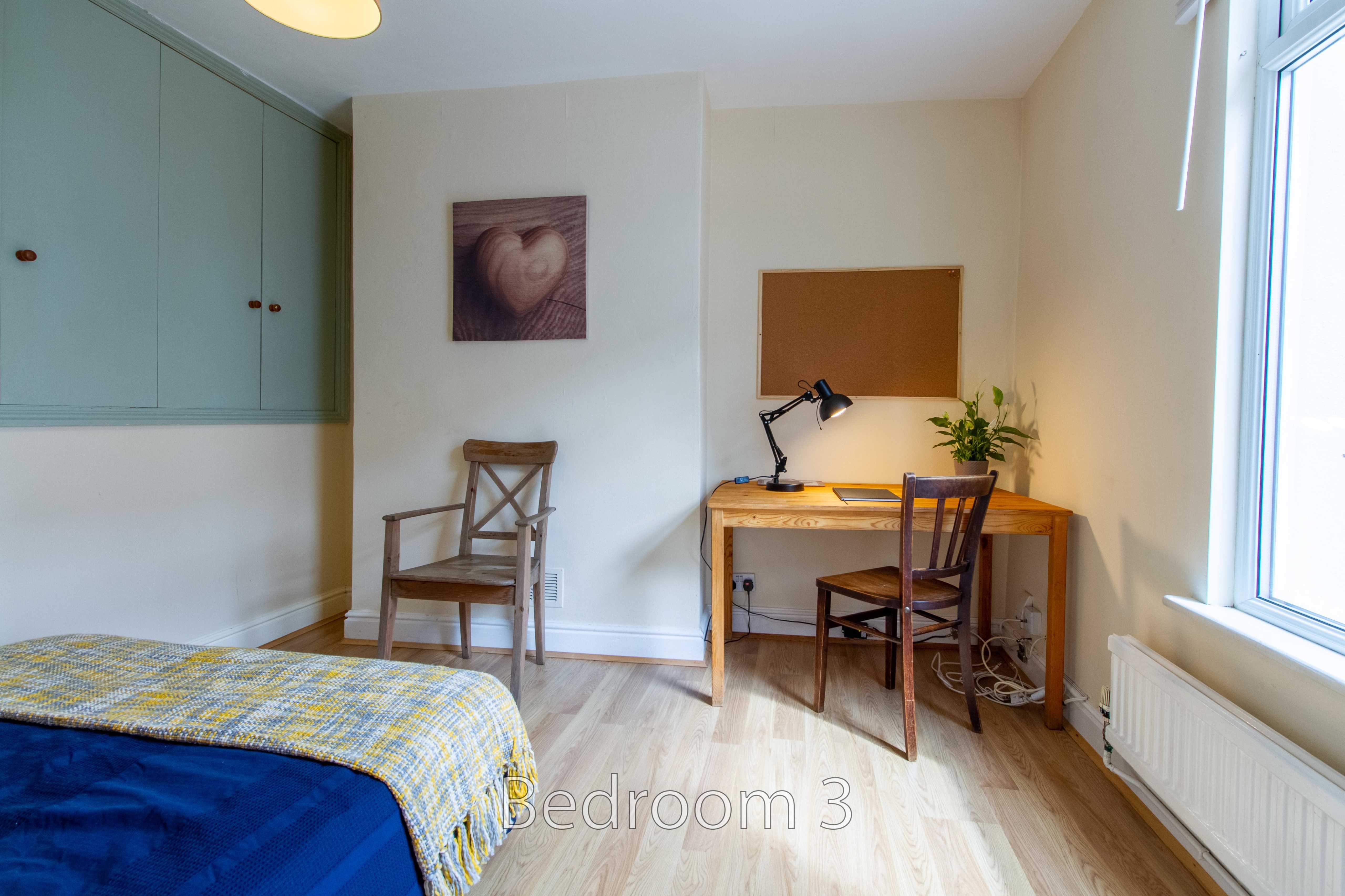 A bedroom with a bed, desk, chairs, built-in storage, a radiator and a large window.