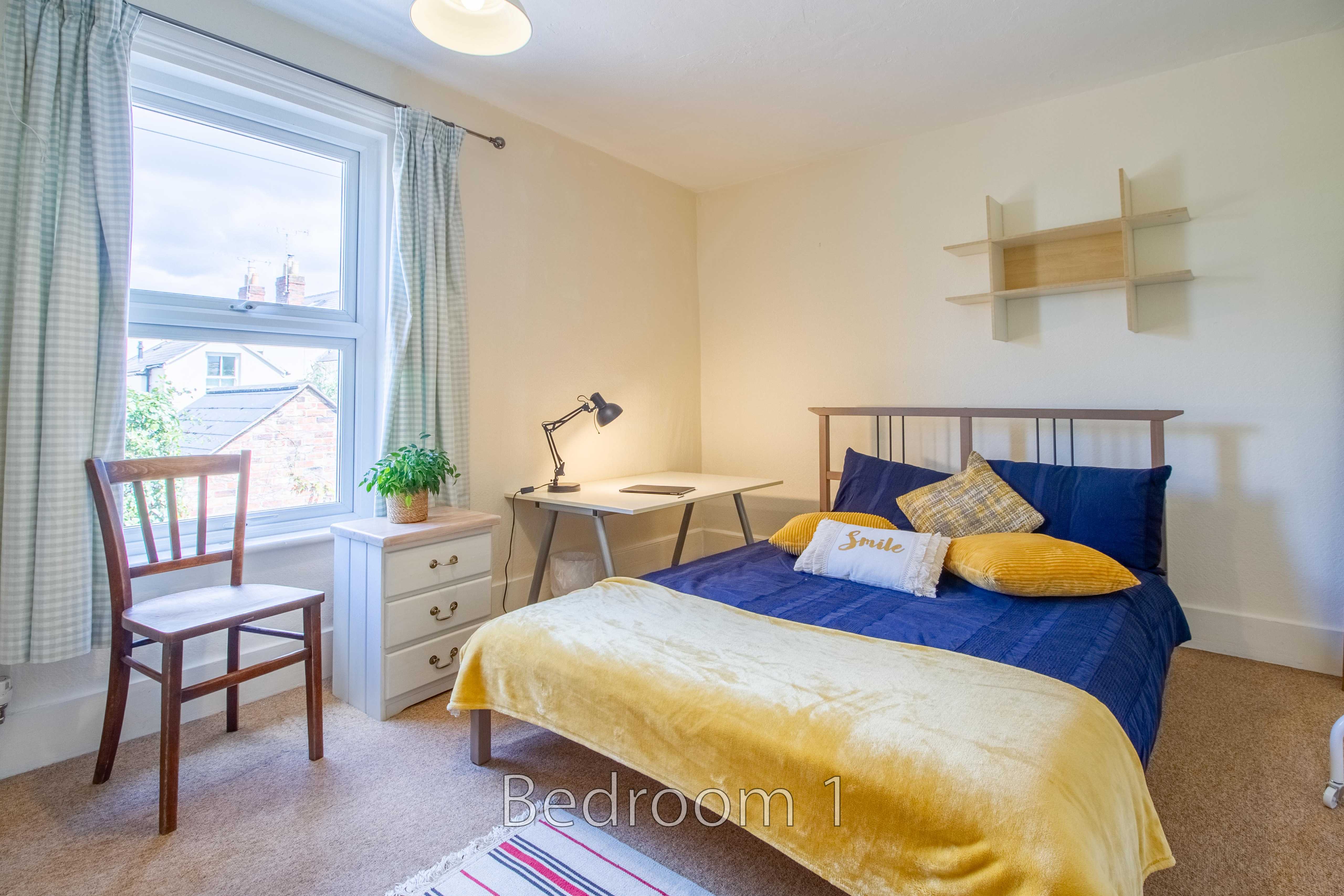 A furnished bedroom with a bed, desk, chair, chest of drawers, window and wall shelves.