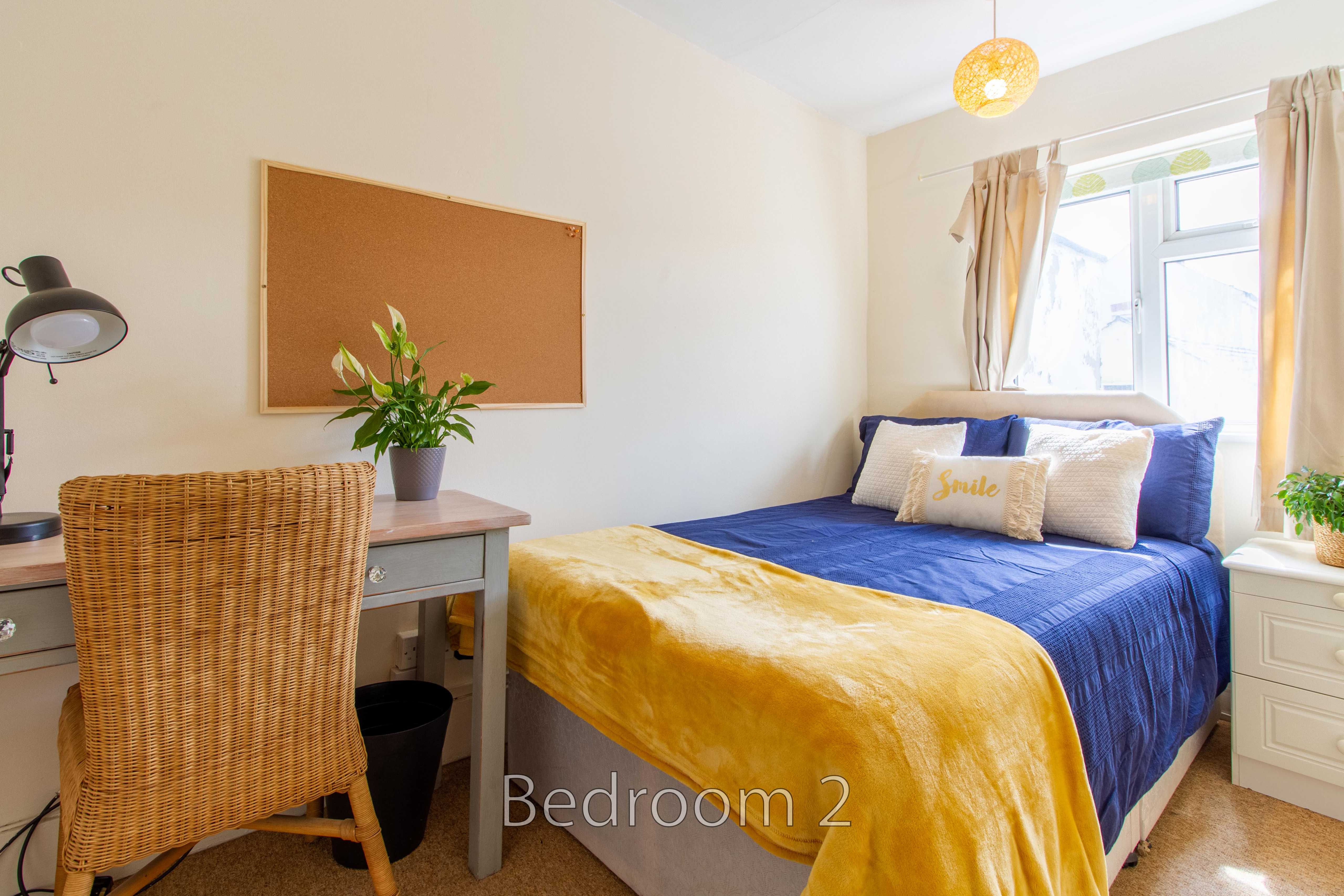 A furnished bedroom with a bed, desk, chair, bedside cabinet, window and yellow-and-blue bedding.