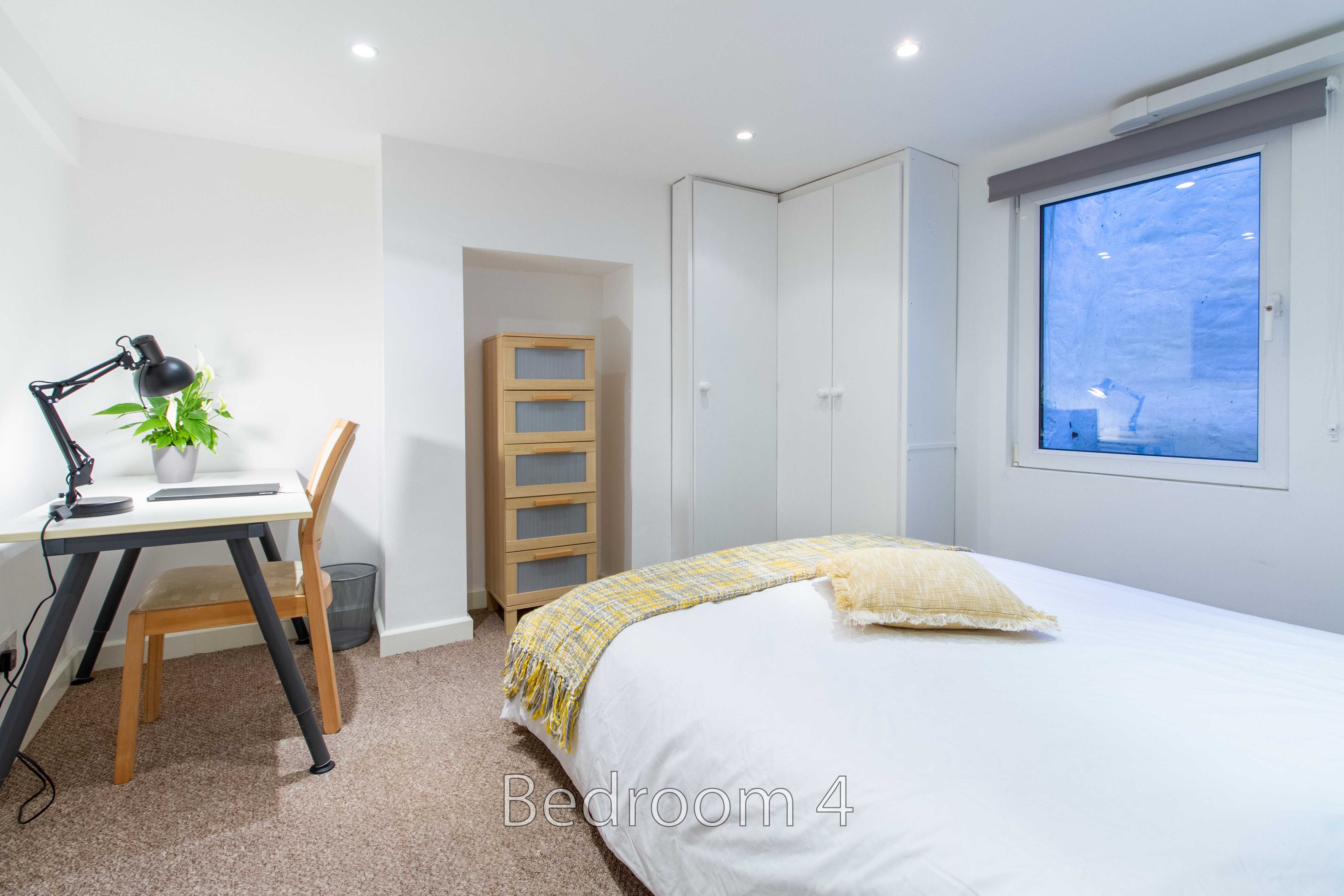 A bright bedroom with a double bed, desk, chair, wardrobe, window and bedside furnishings.
