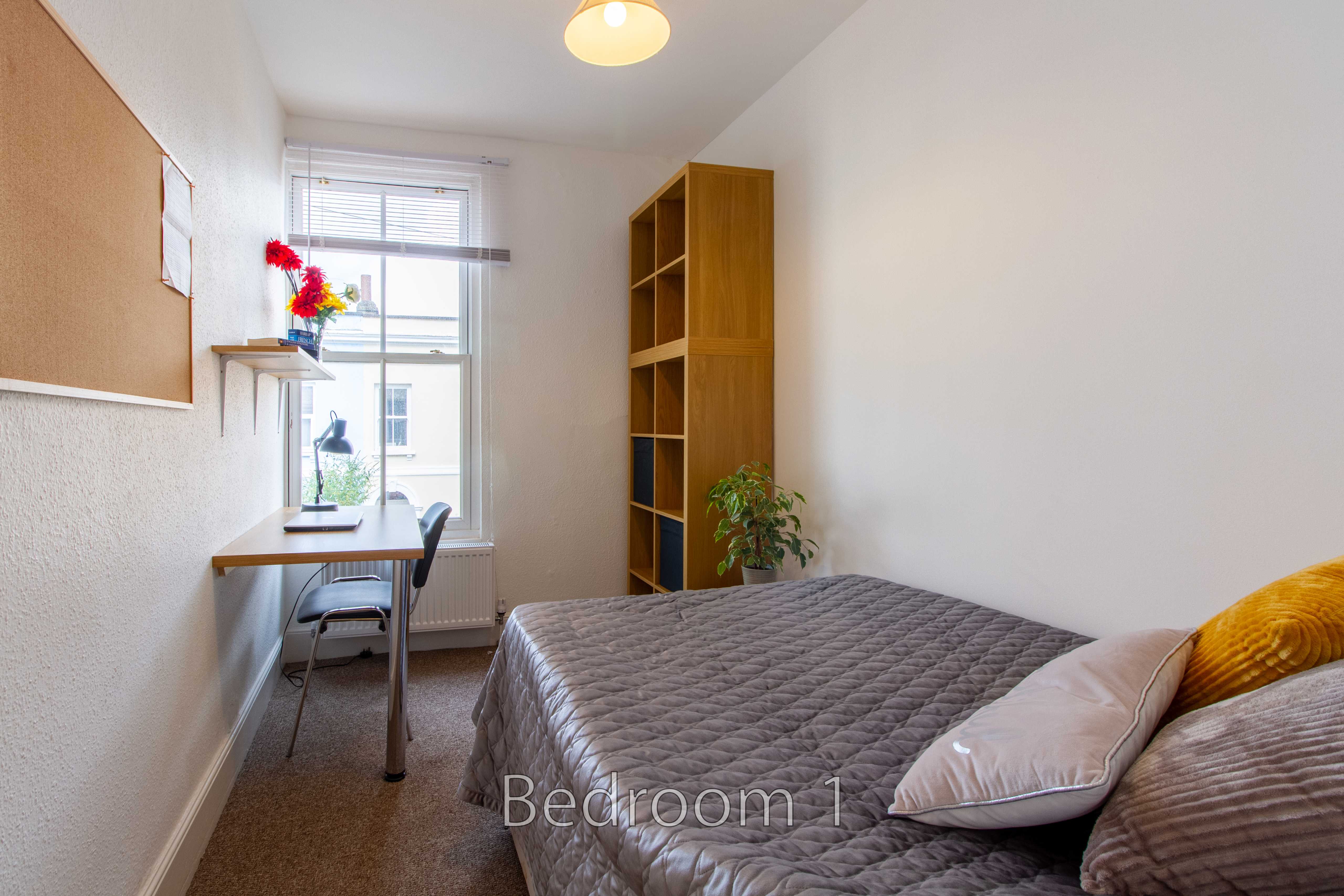 A furnished bedroom with a double bed, desk, chair, shelving unit, window and potted plants.