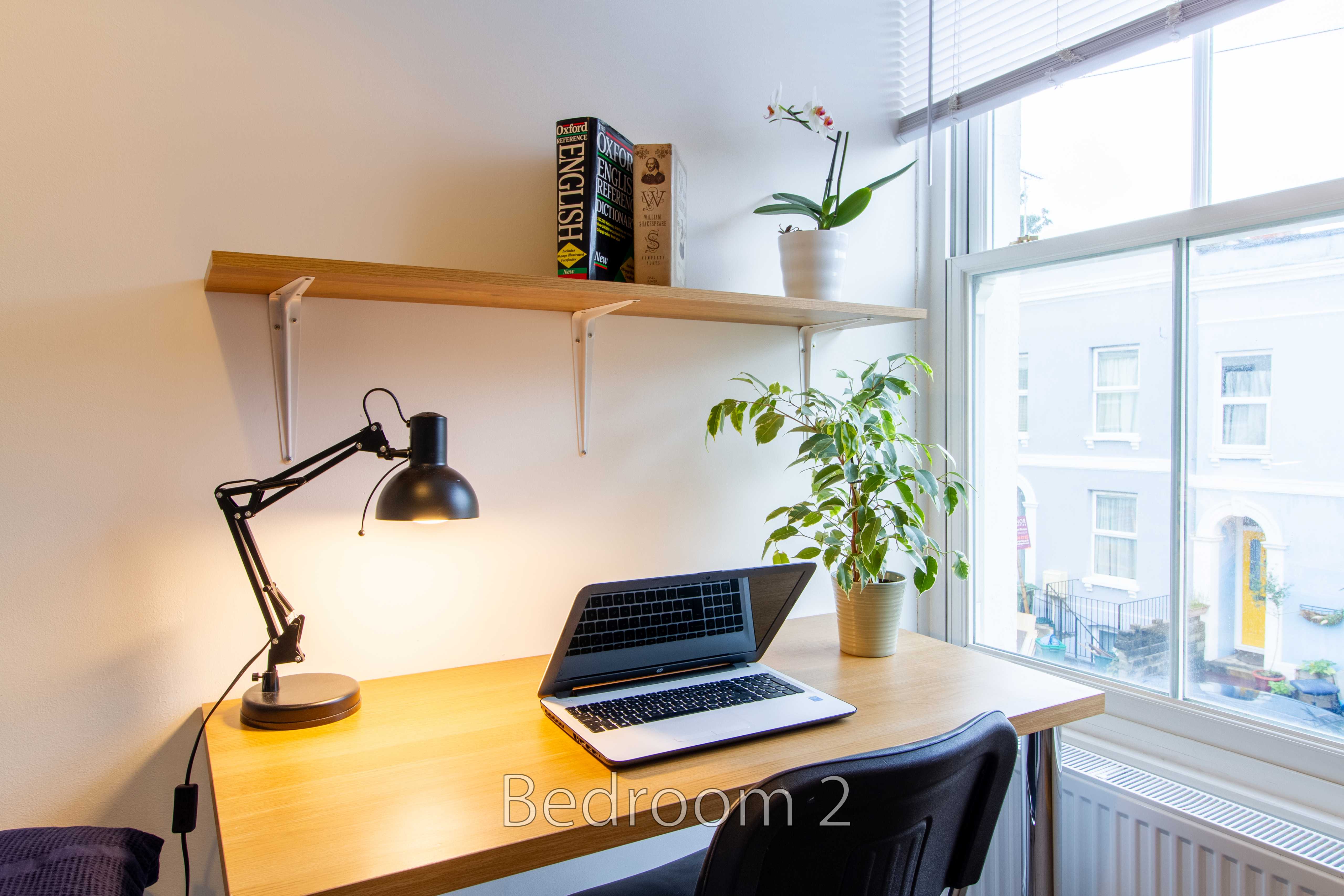 Bedroom with a desk, open laptop, lamp, shelves, plants and a large window.