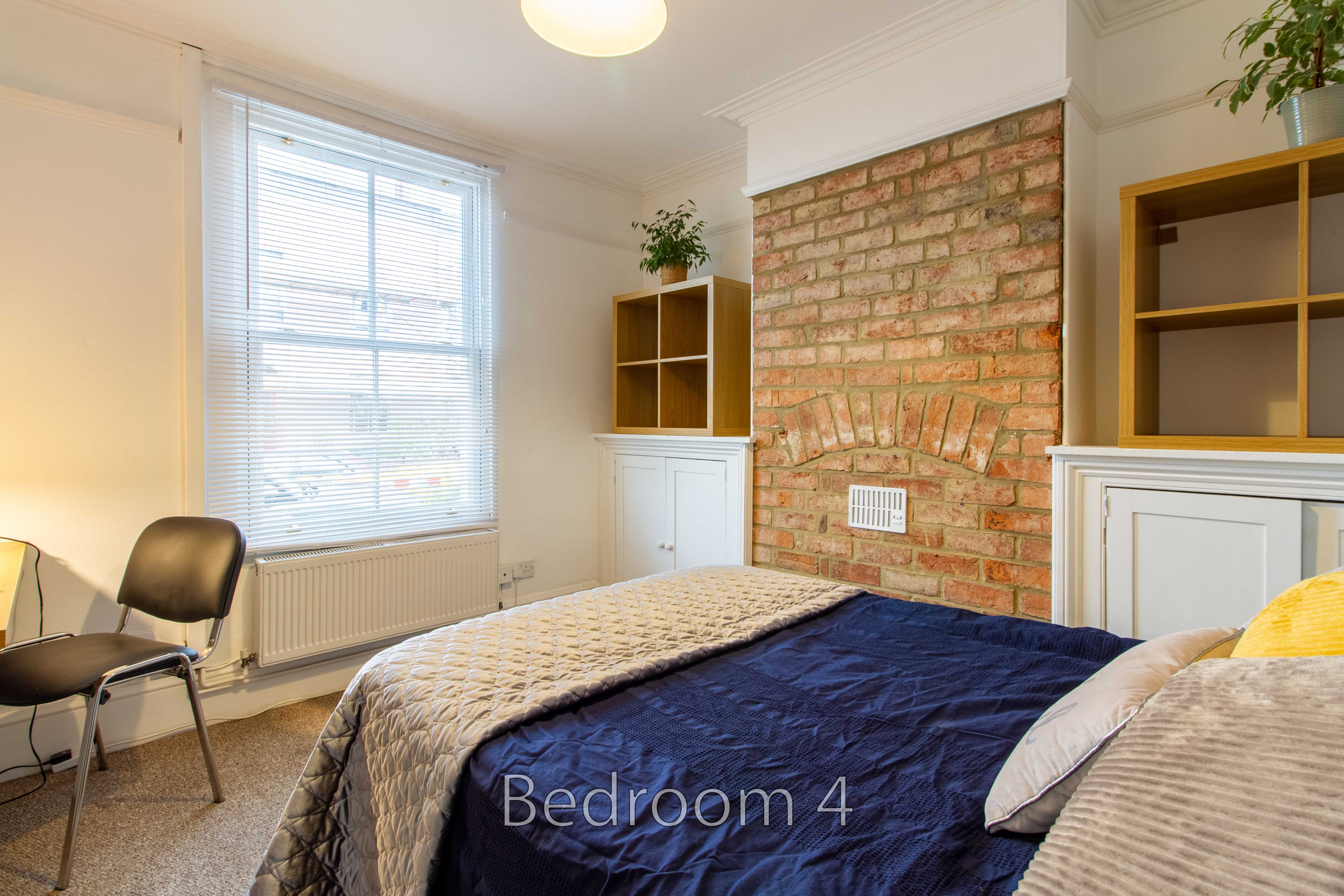 A bedroom with two beds, a brick fireplace, built-in shelving, a window with blinds, and a radiator.