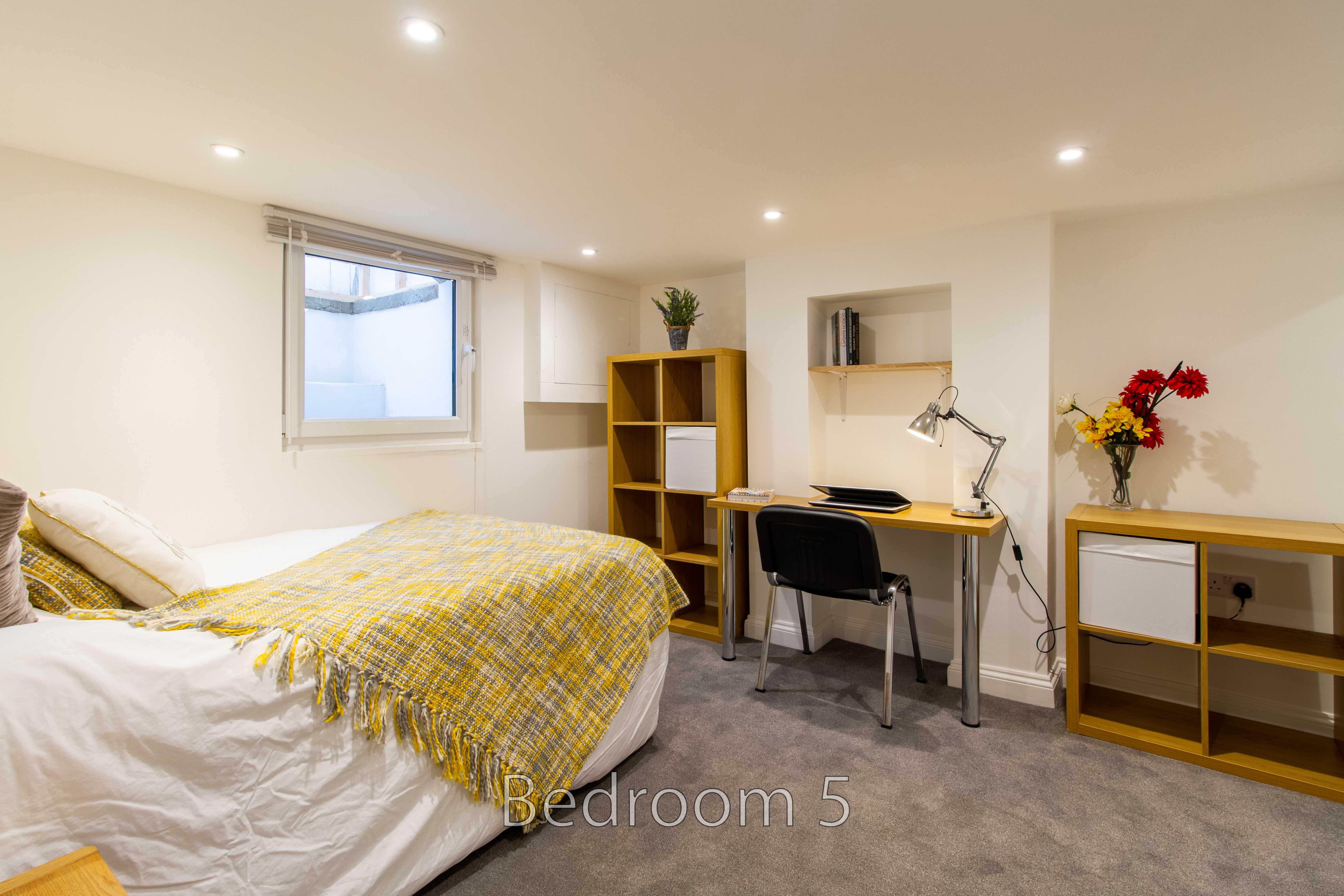 A furnished bedroom with a bed, desk and chair, shelving, a window and a vase of flowers.