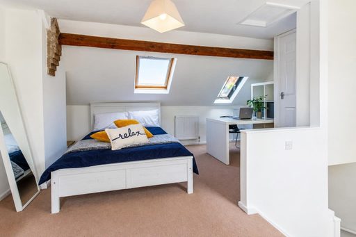 A furnished attic bedroom with a double bed, skylights, exposed wooden beam, desk and wardrobe.