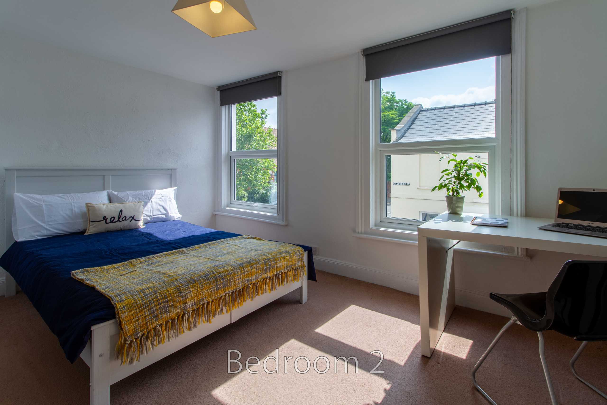 A bright bedroom with a double bed, two windows, a desk, chair and potted plant.
