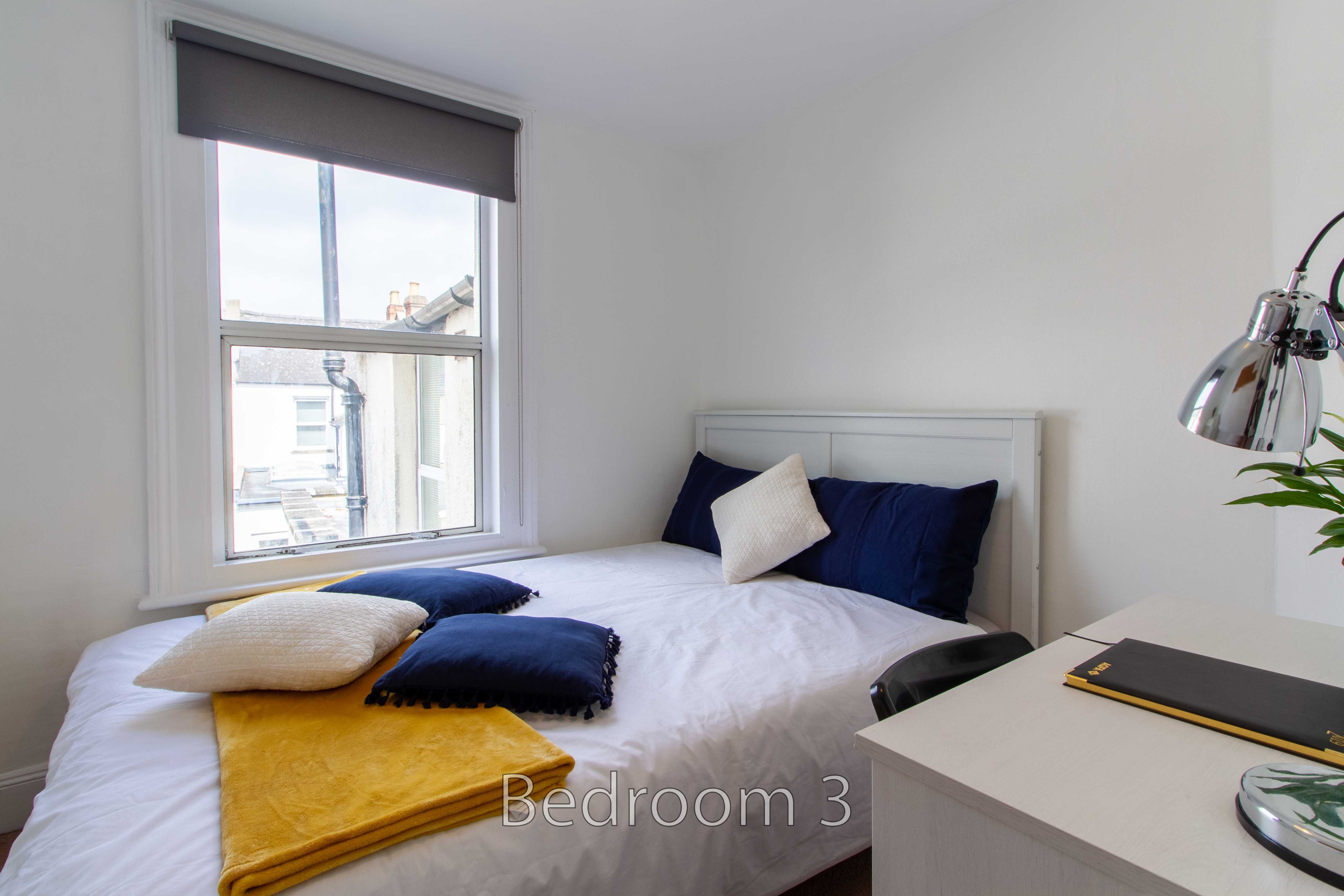 A bedroom with a double bed, blue and white pillows, a window, a desk, and a lamp.
