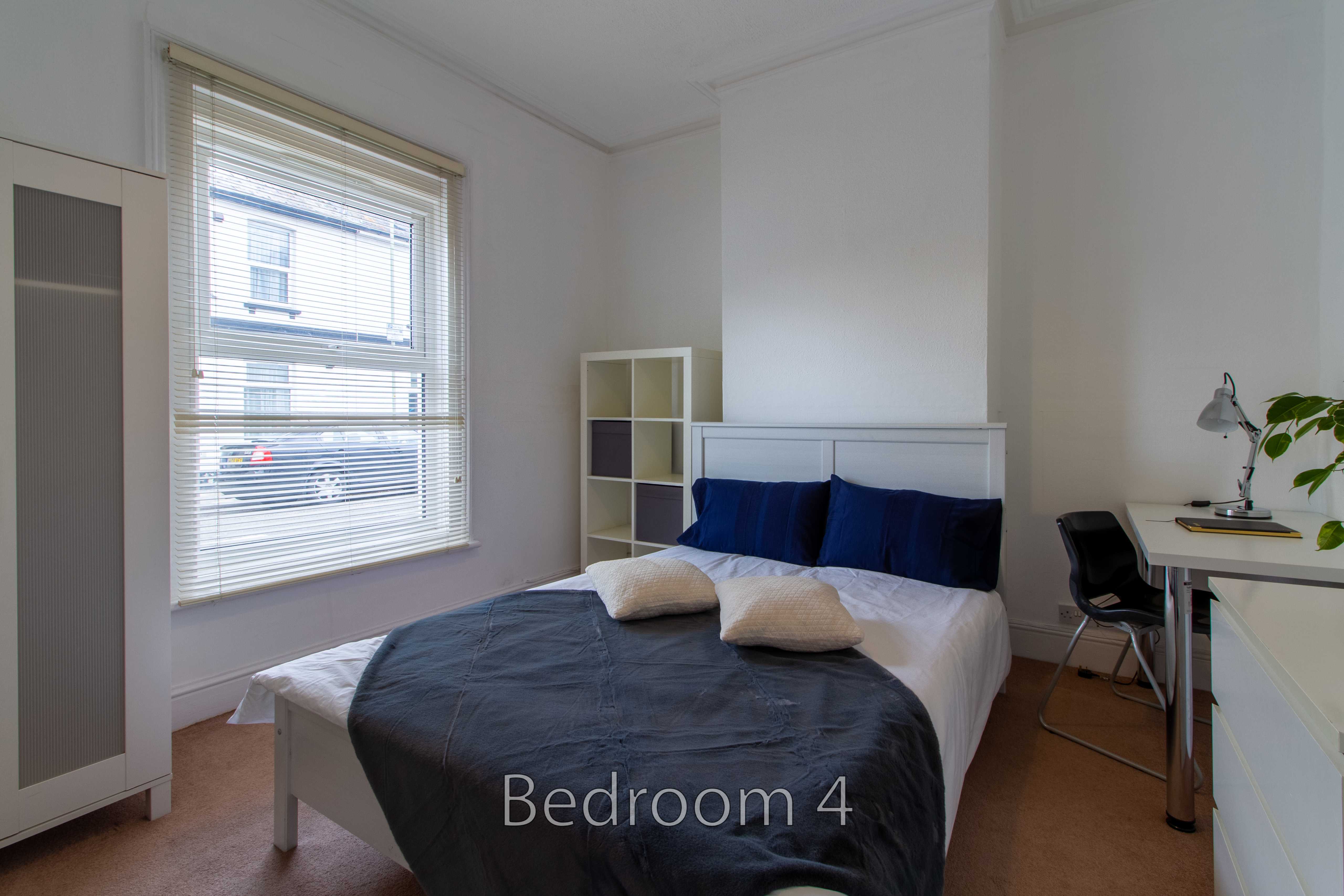 A furnished bedroom with a double bed, wardrobe, shelving unit, desk and window with blinds.