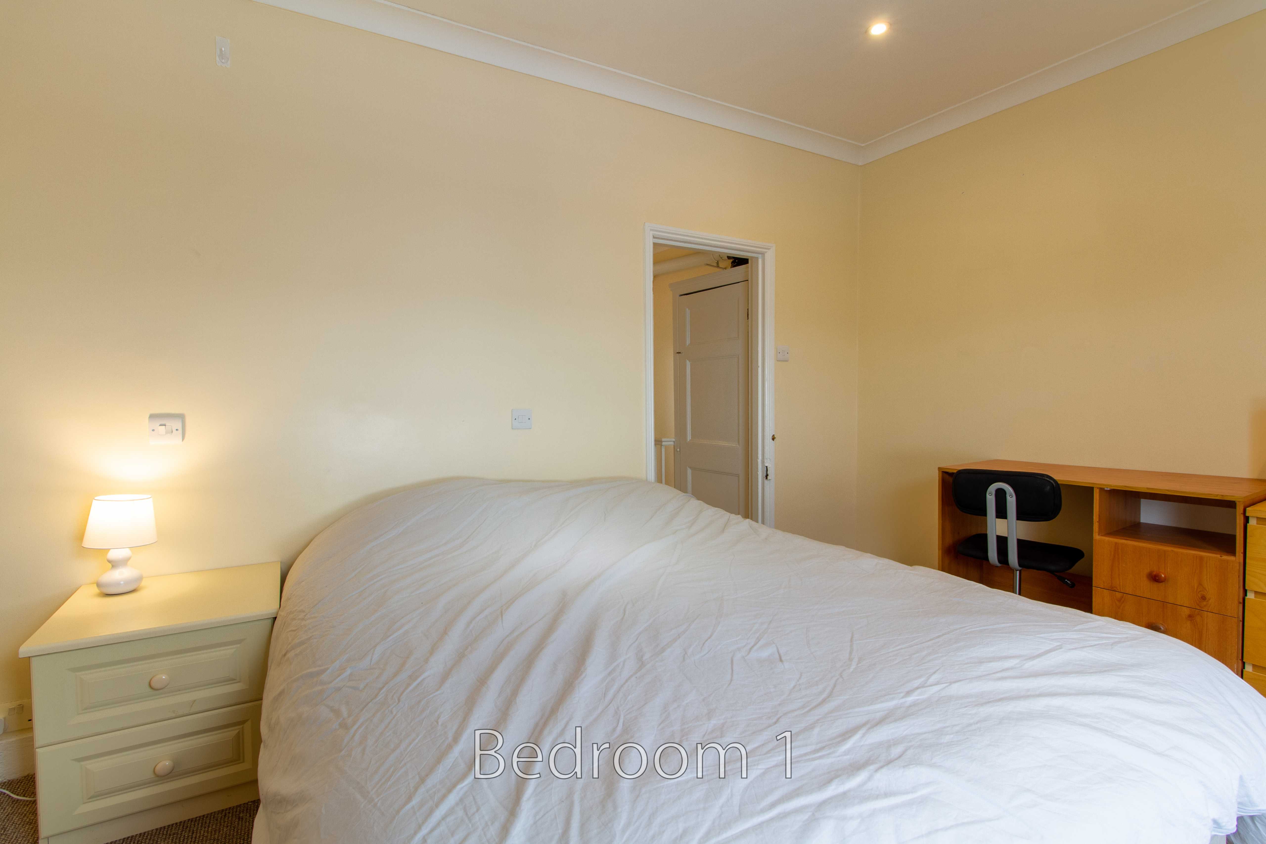 A furnished bedroom with a double bed, bedside table and lamp, desk, chair and wardrobe.