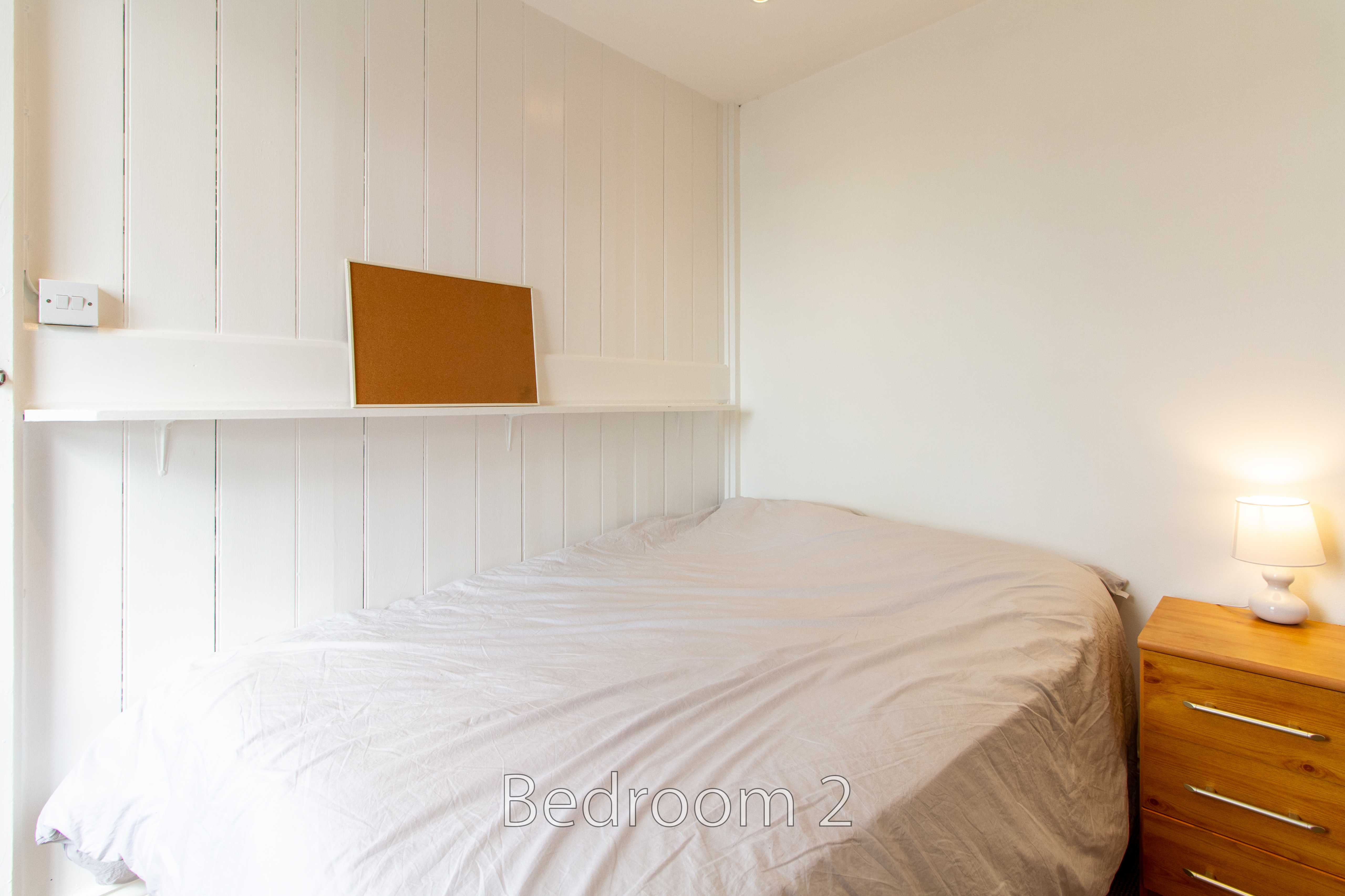 A bedroom with a double bed, white panelled walls, a shelf, and a wooden bedside chest.
