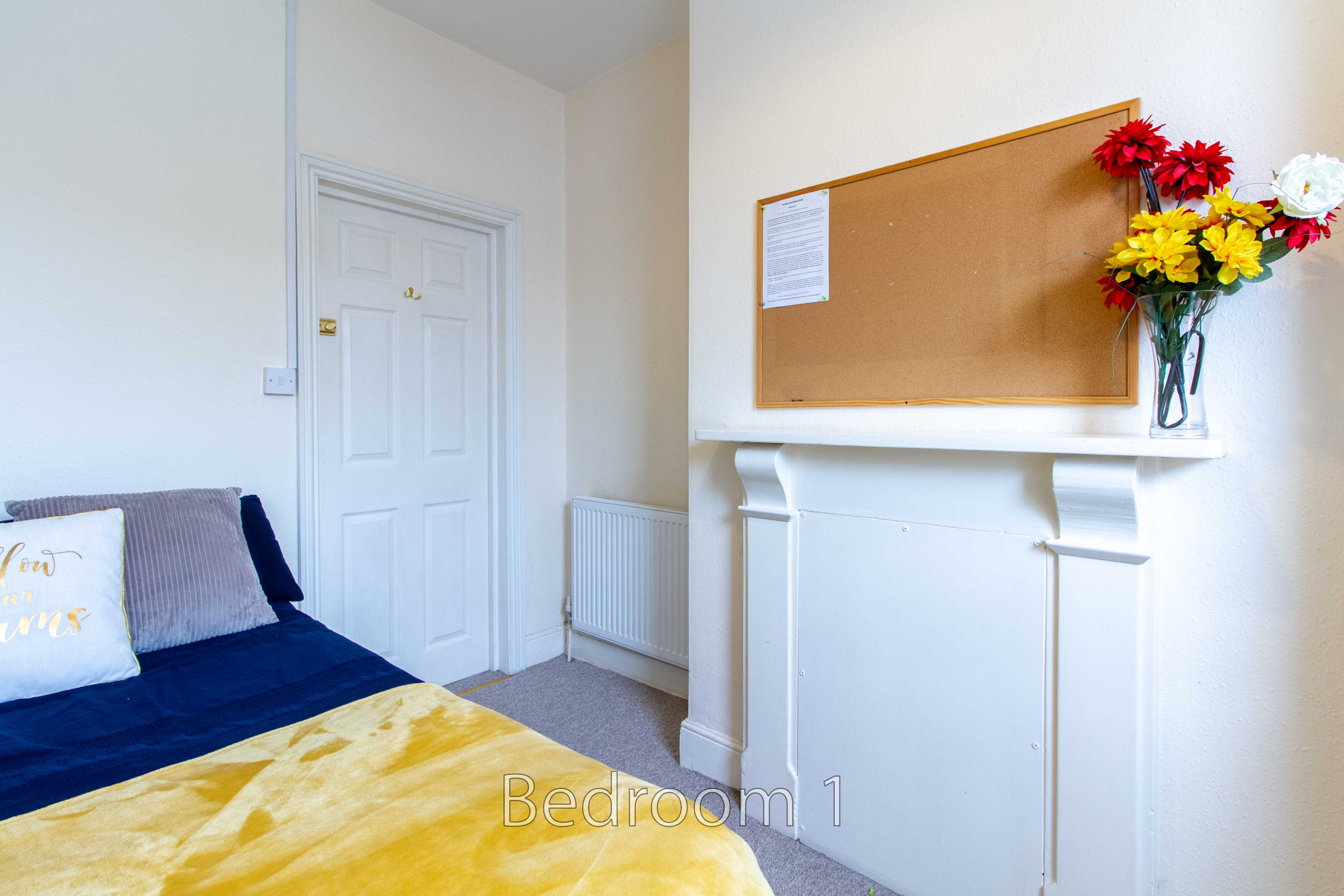 A bedroom with a bed, white door, radiator, fireplace surround and wall-mounted noticeboard.