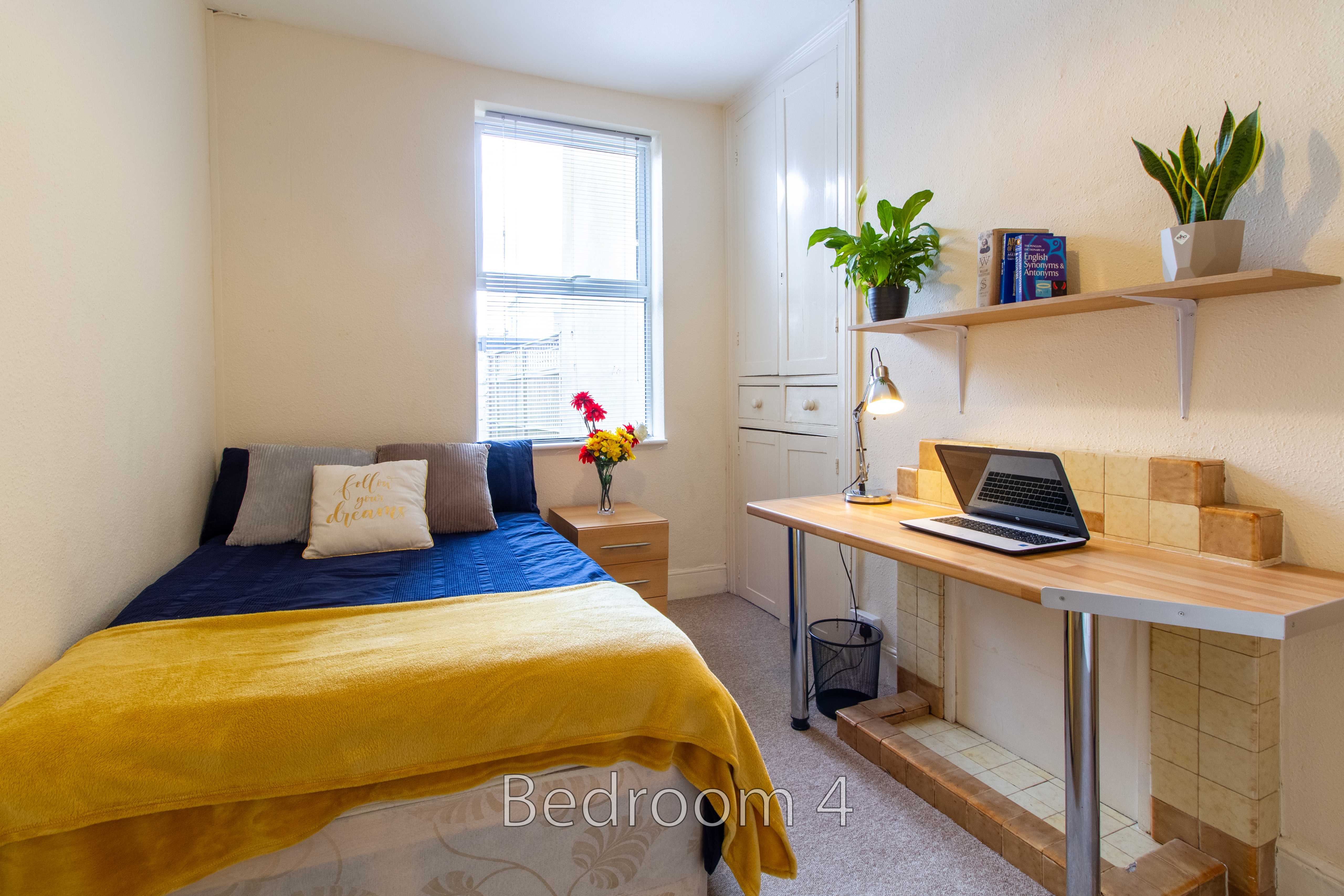 A furnished bedroom with a bed, desk, laptop, window, shelves and potted plants.