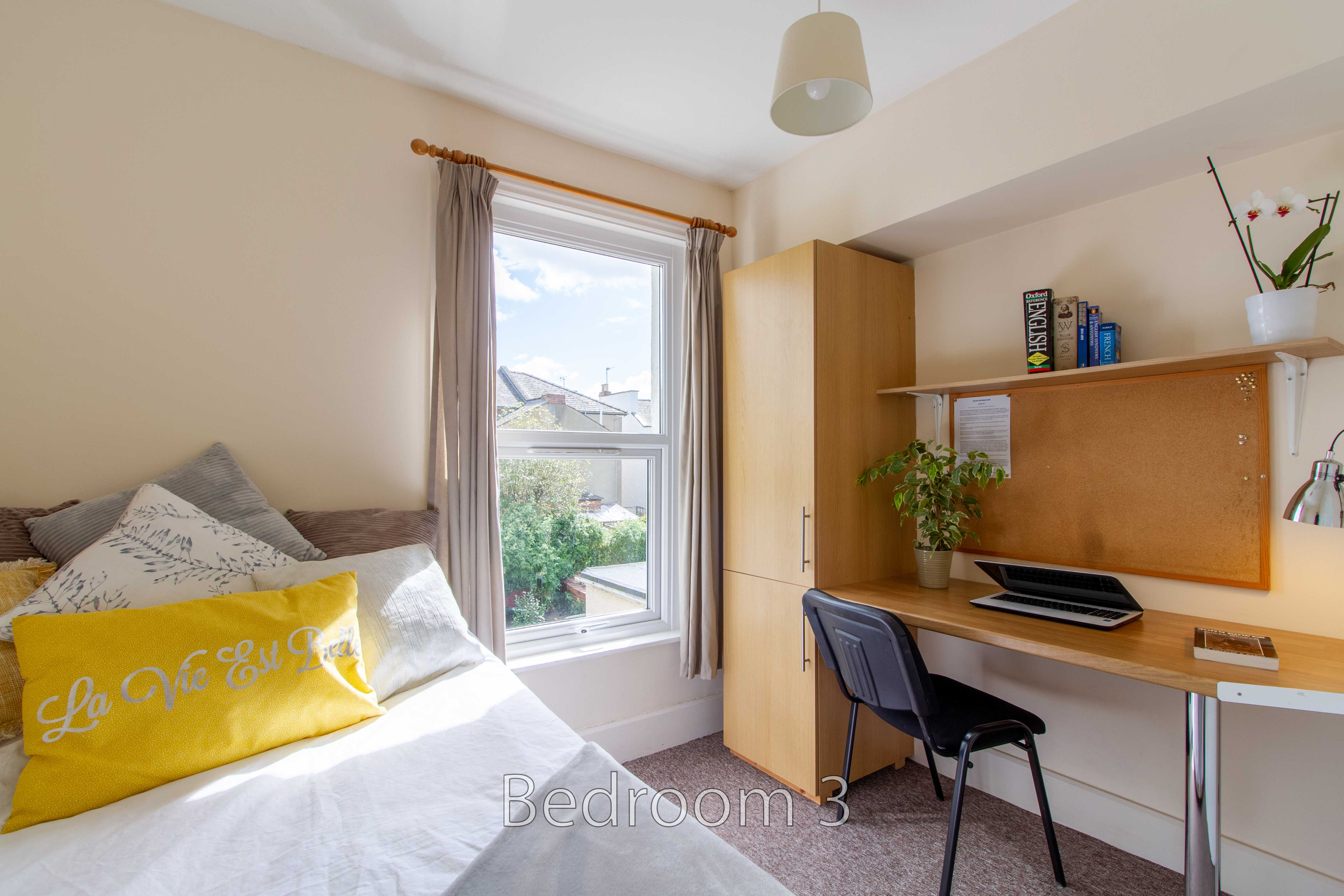 A furnished bedroom with a single bed, desk, chair, wardrobe, window and plants.