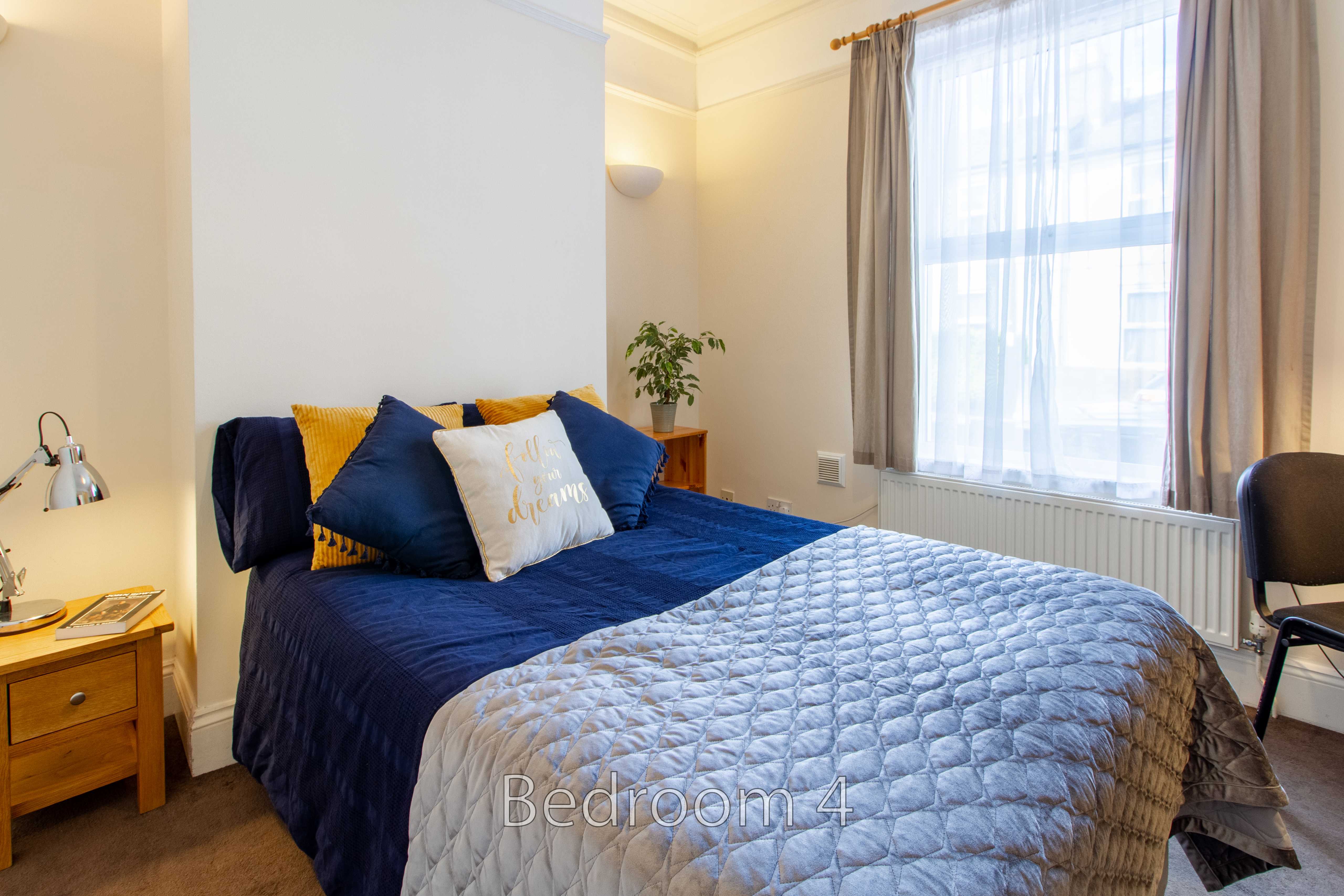 A bedroom with a double bed, blue and grey bedding, bedside tables, curtains and a large window.