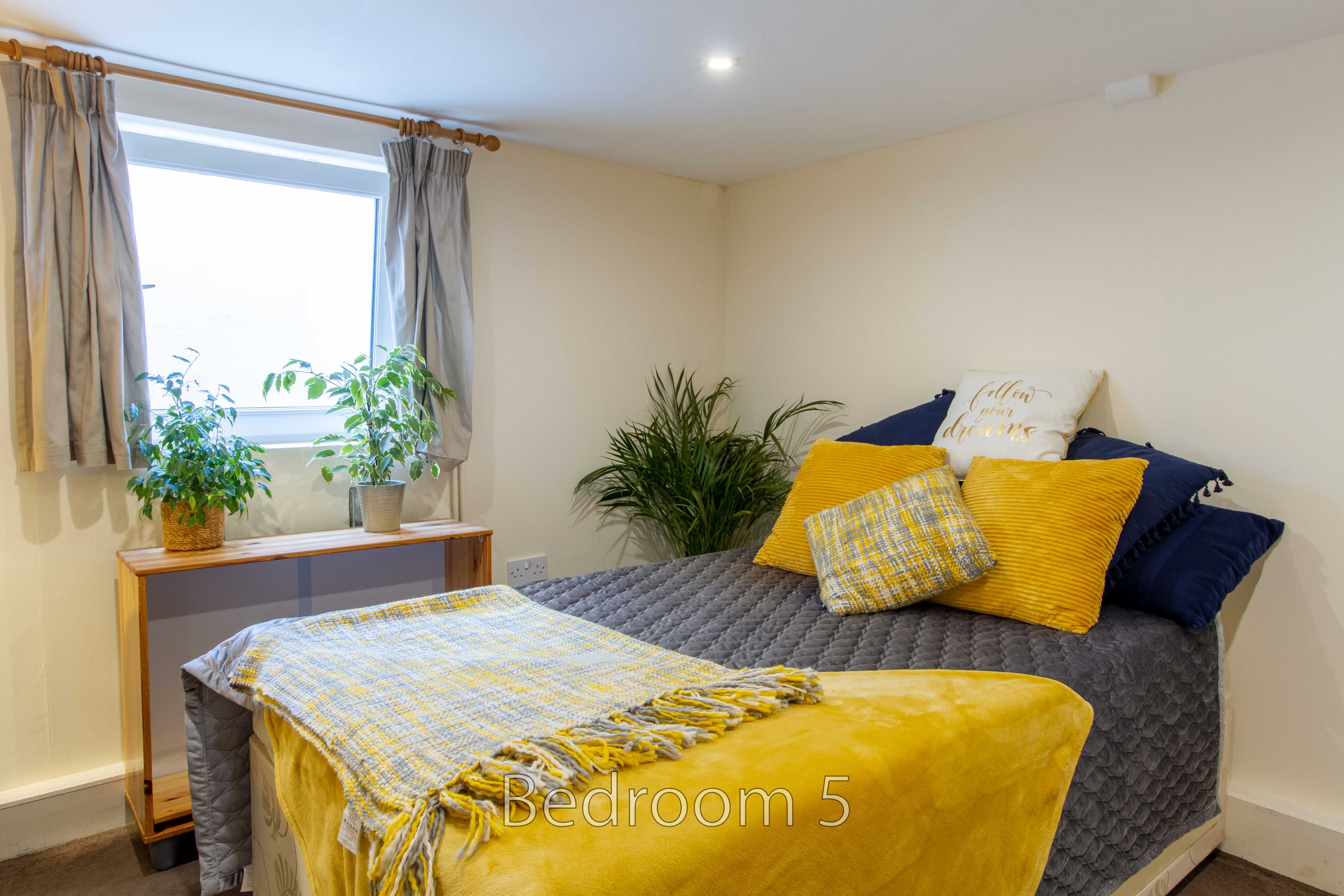 A furnished bedroom with a double bed, yellow and blue bedding, a window, curtains, plants and a small table.